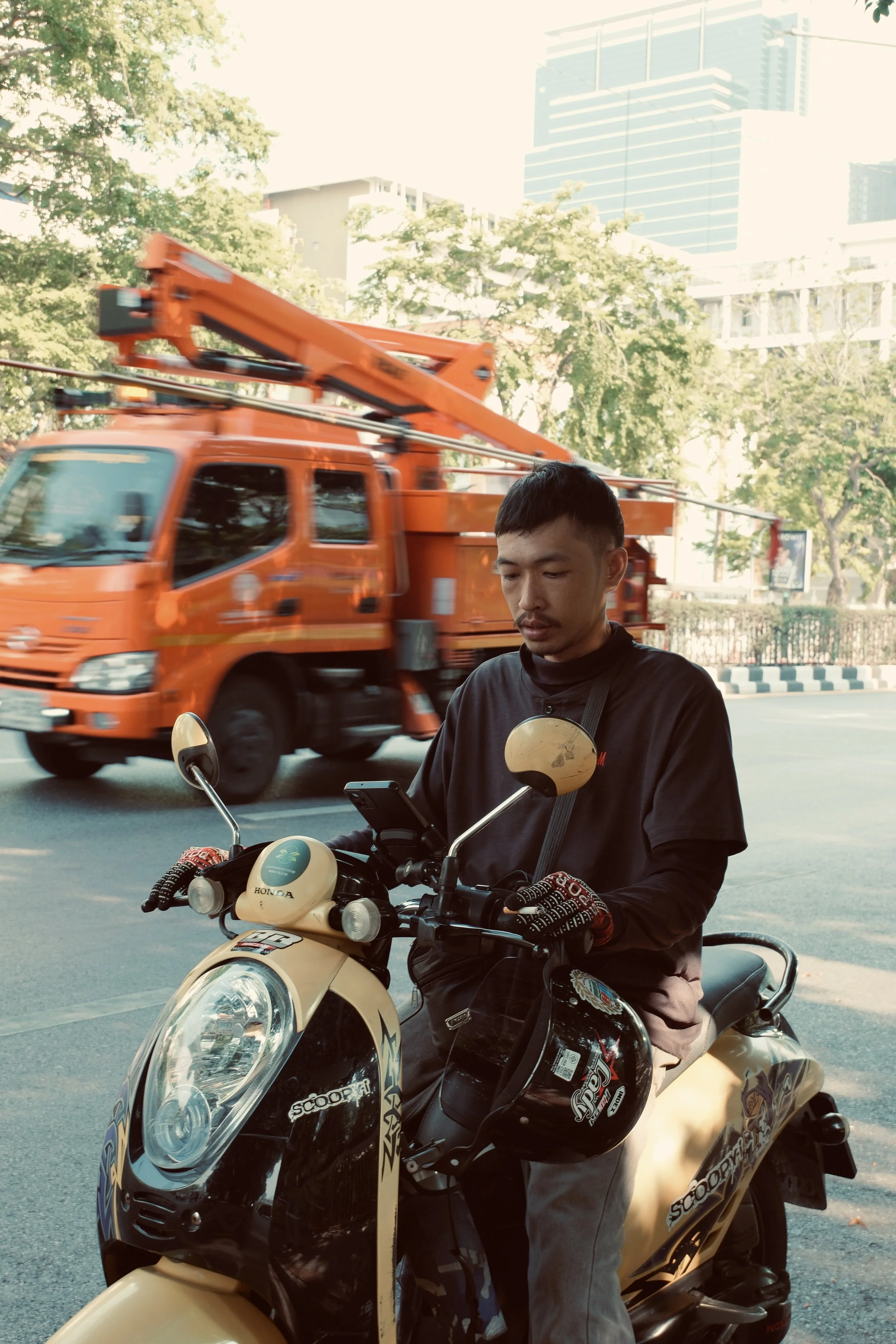 A man on a beige scooter using a smartphone on a city street with a red fire truck passing by in the background, trees, and tall buildings.