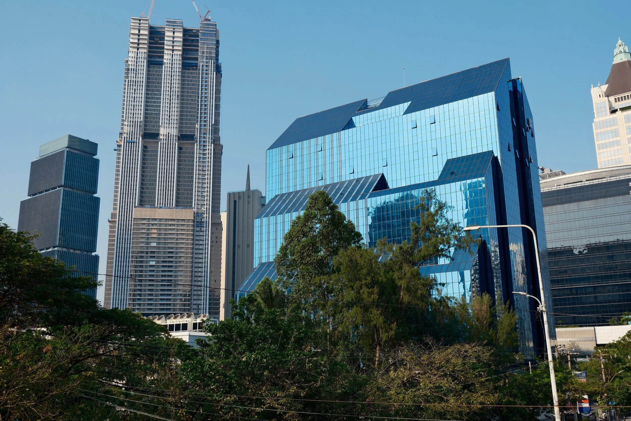 City skyline with modern high-rise buildings, one with reflective blue glass windows, and green trees in the foreground under a clear blue sky.