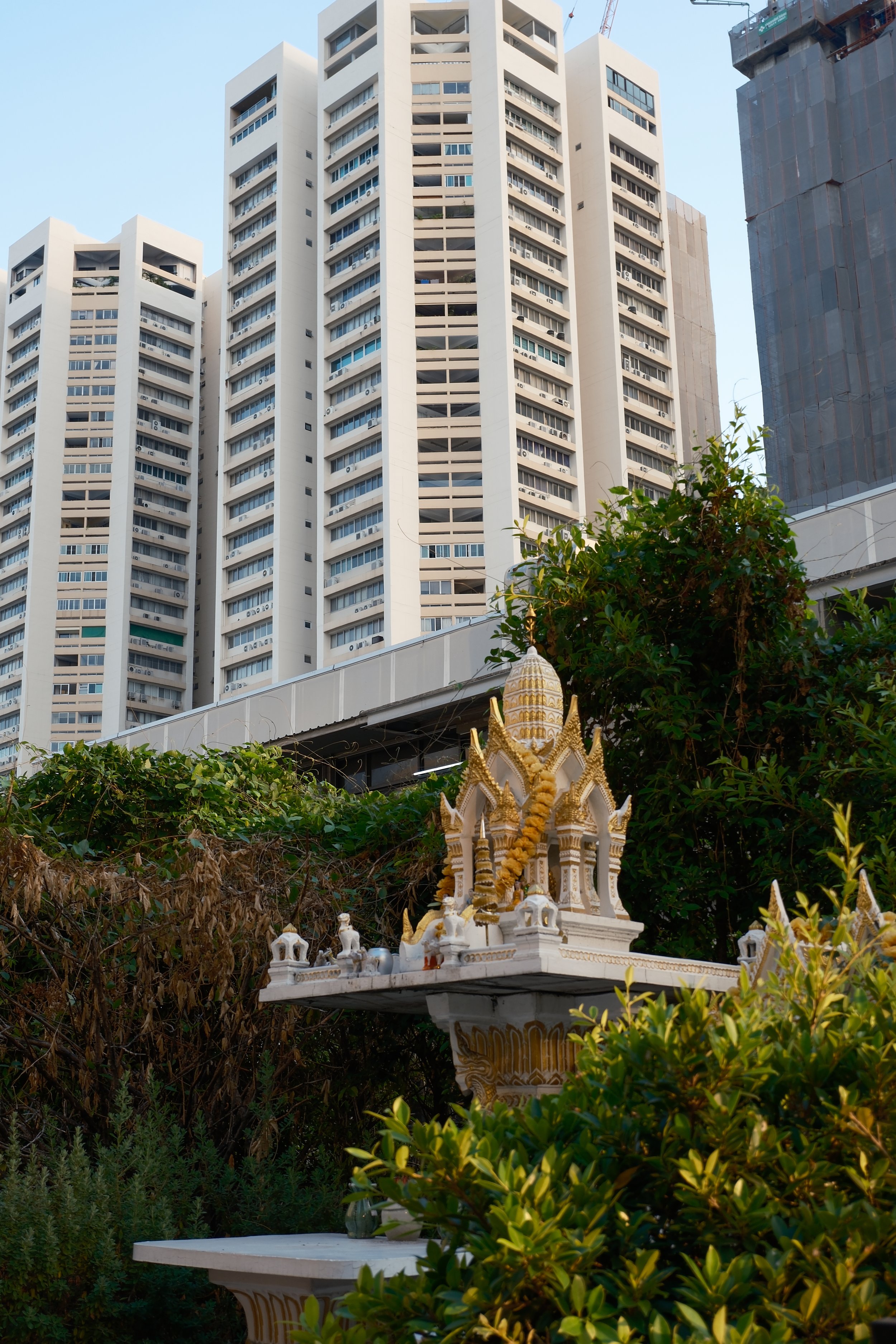 A small traditional shrine with intricate detailing and gold accents, set amidst greenery and bushes, with tall modern apartment buildings in the background under a clear blue sky.