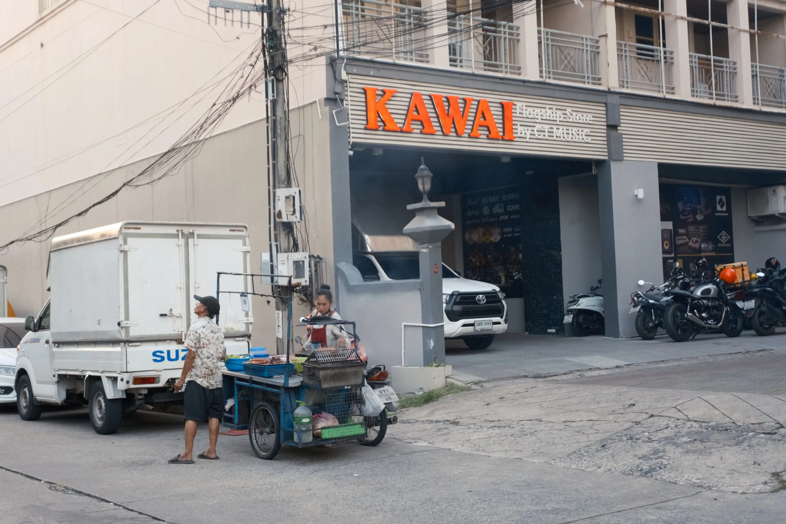 Street view with a small food cart and a vendor in front of a building with signs for KAWAI and a motorcycle shop.