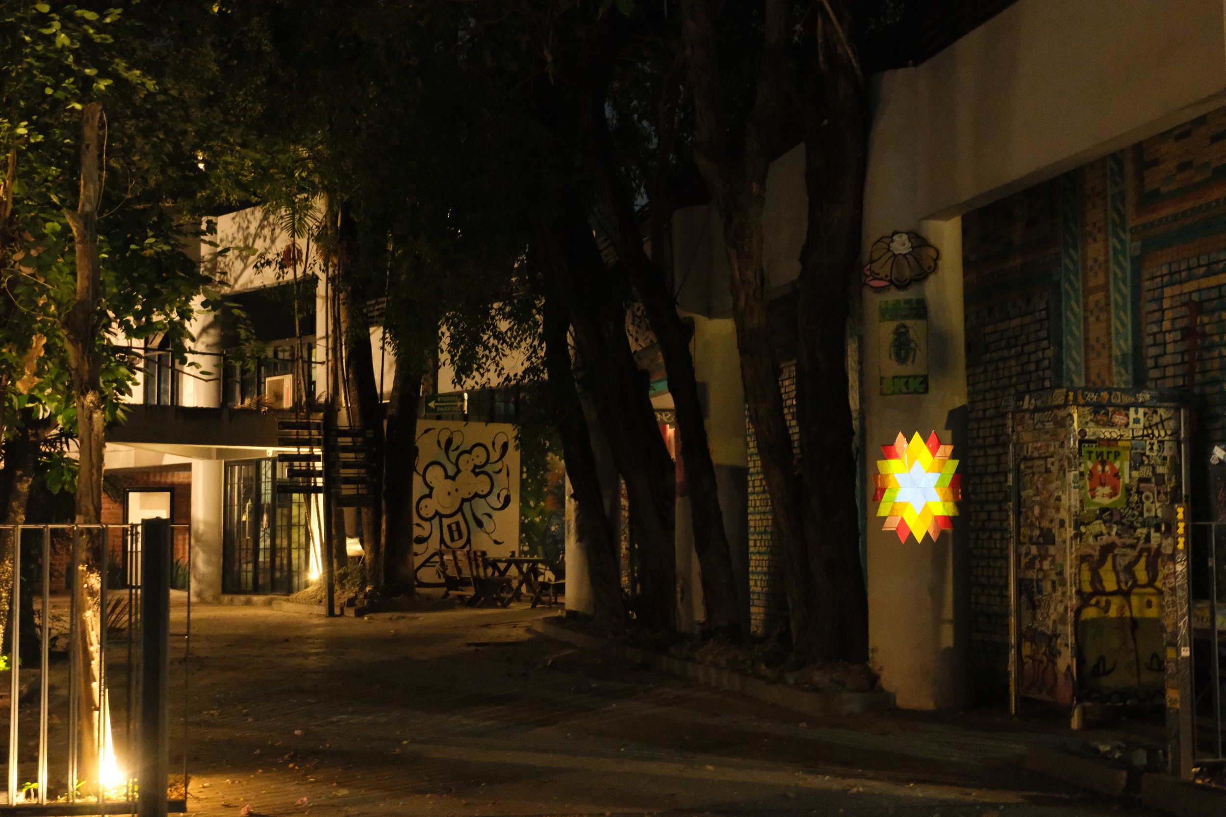 A dimly lit outdoor scene at night featuring trees, a wall with colorful murals, and a bright, multi-colored star-shaped light fixture.