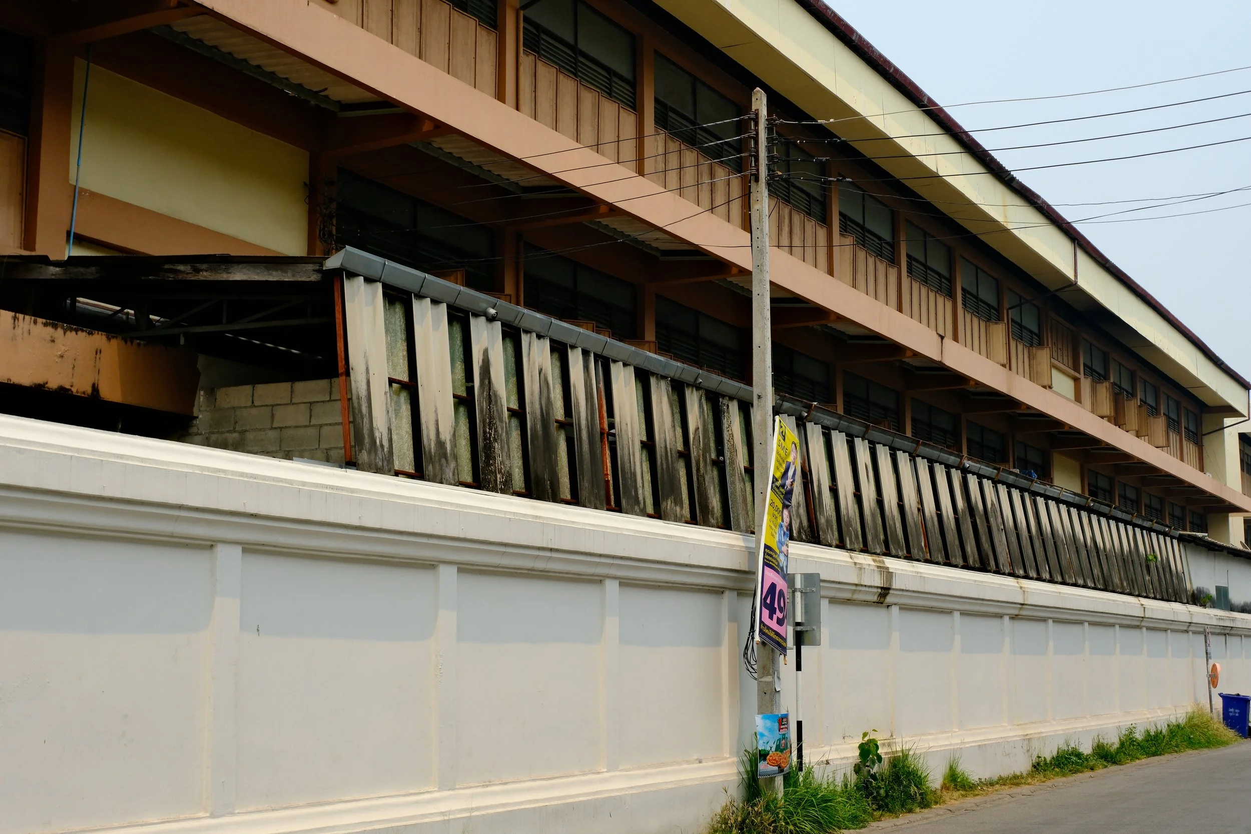 A multi-story building with a worn wooden balcony and a white wall below. A utility pole with a poster and some plants in front of it are visible in the sidewalk.