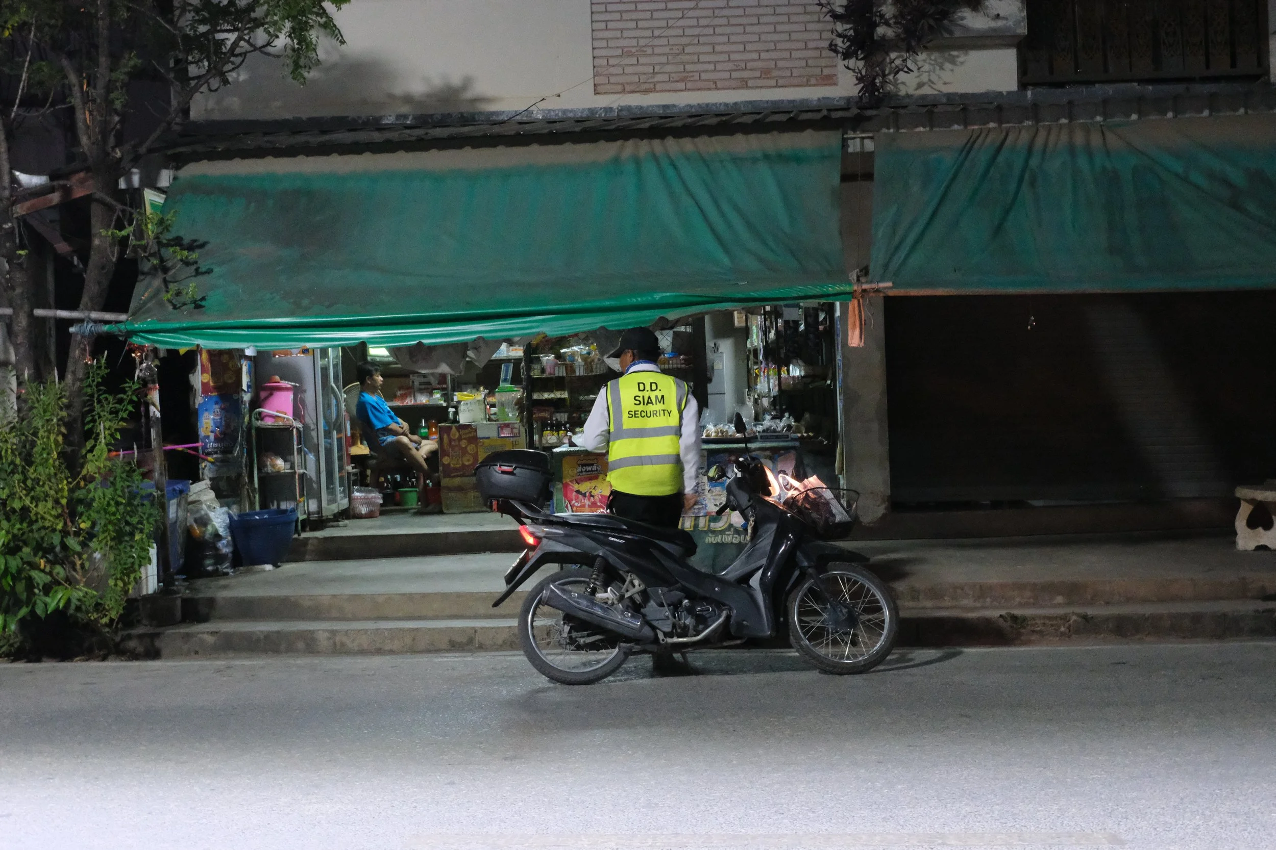 A security guard in a yellow vest with 'D.D. SIAM SECURITY' on the back standing next to a black motorcycle in front of a small shop with a green awning, on a street at night.