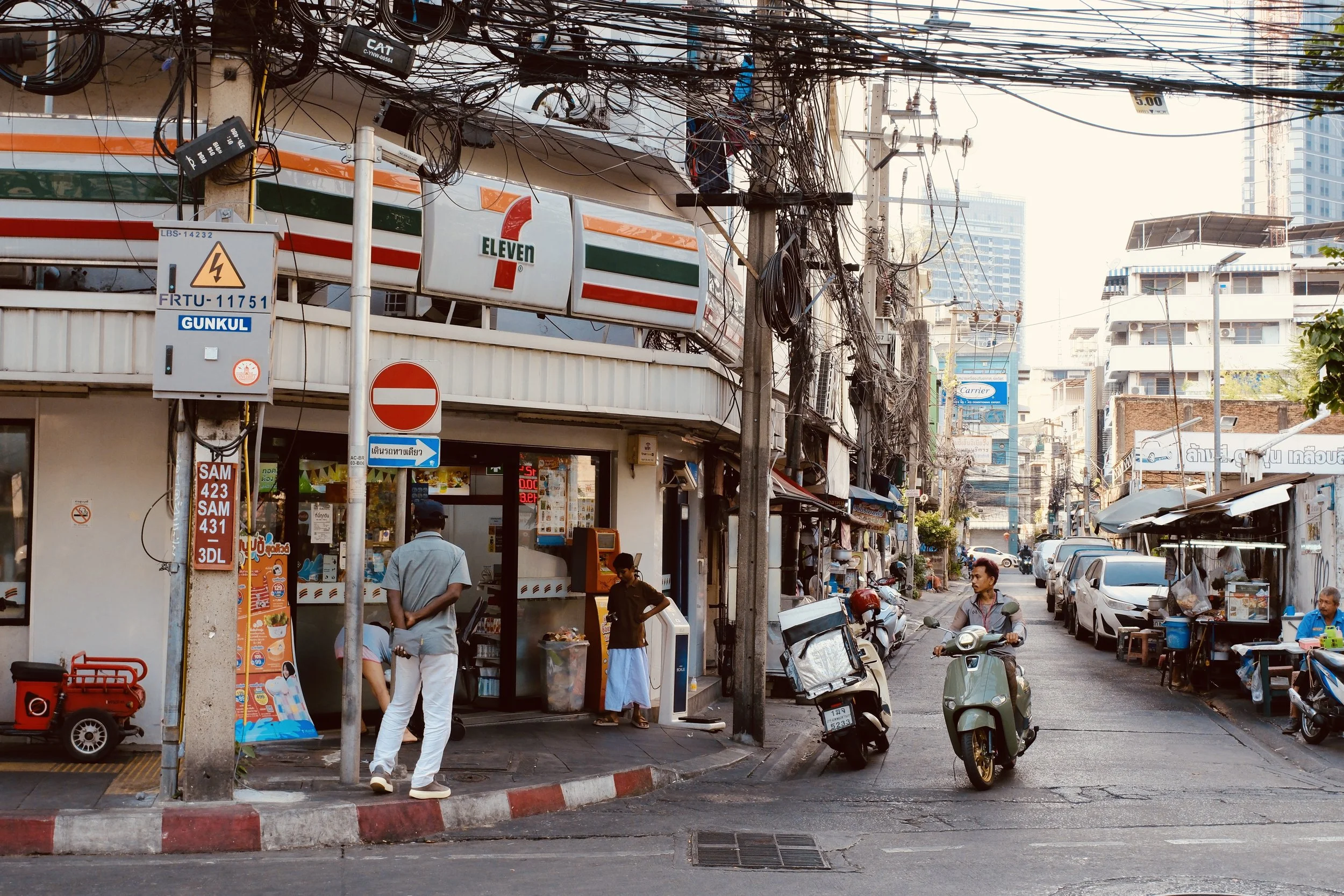 Street scene in an urban area with a 7-Eleven convenience store, motorbikes parked on the sidewalk, a person riding a scooter, and electrical wires overhead.