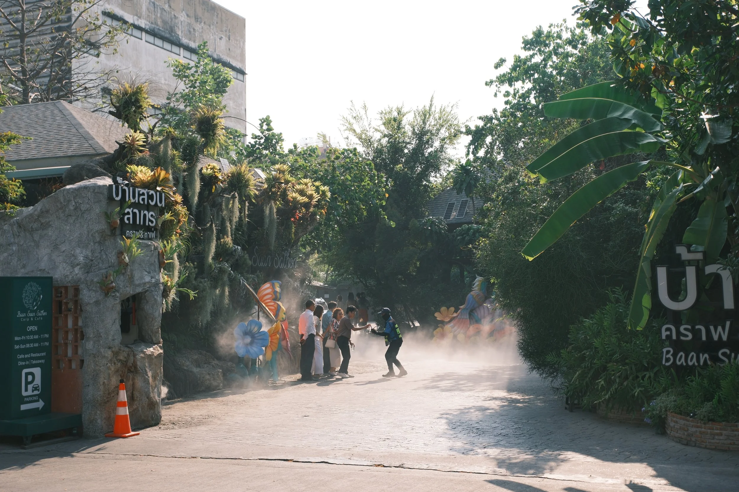 People standing in front of the entrance of Baan Suan Satar garden, with colorful decorations and greenery, some visitors shaking hands or greeting each other, outdoor setting with trees and a building in the background.