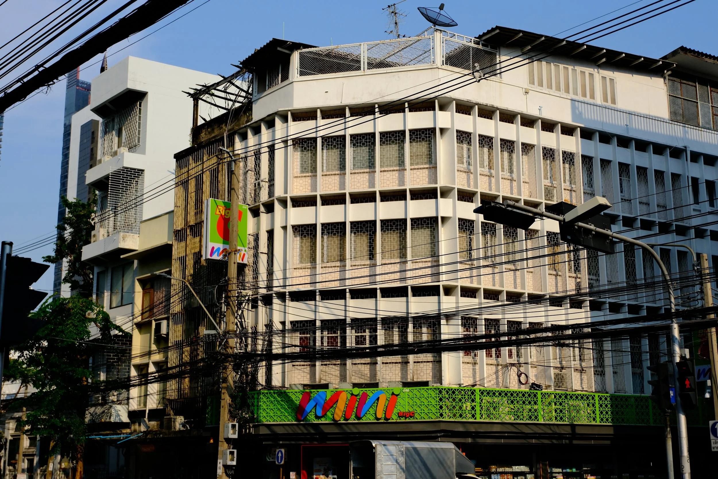 A multi-story building with a green balcony railing, a colorful sign that reads "Mini Big C," and various electrical wires in front.