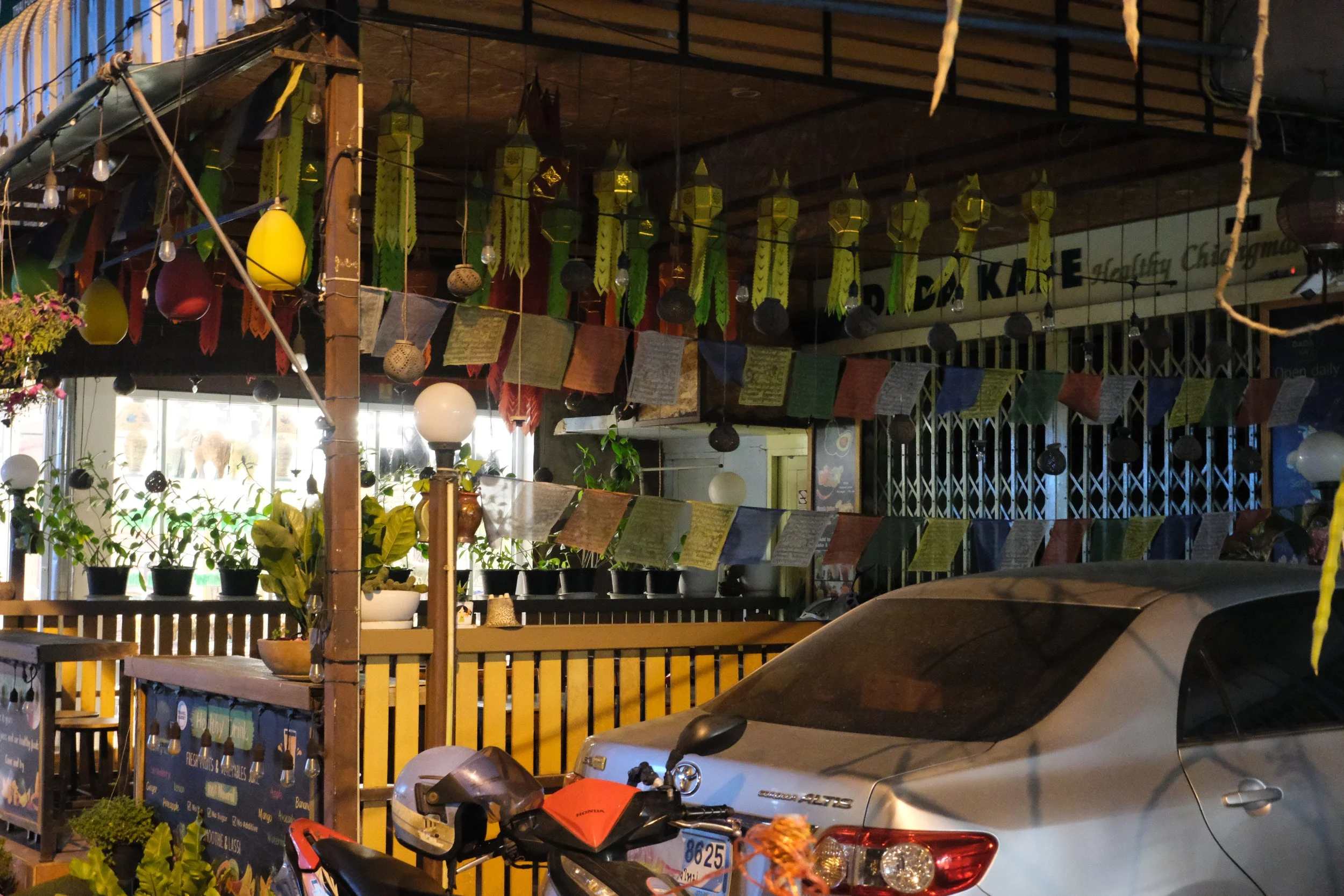 An outdoor seating area of a cafe or restaurant decorated with colorful prayer flags hanging from the ceiling. There are potted plants and lanterns, with a silver car and motorcycle parked in the foreground. The background features large windows and 