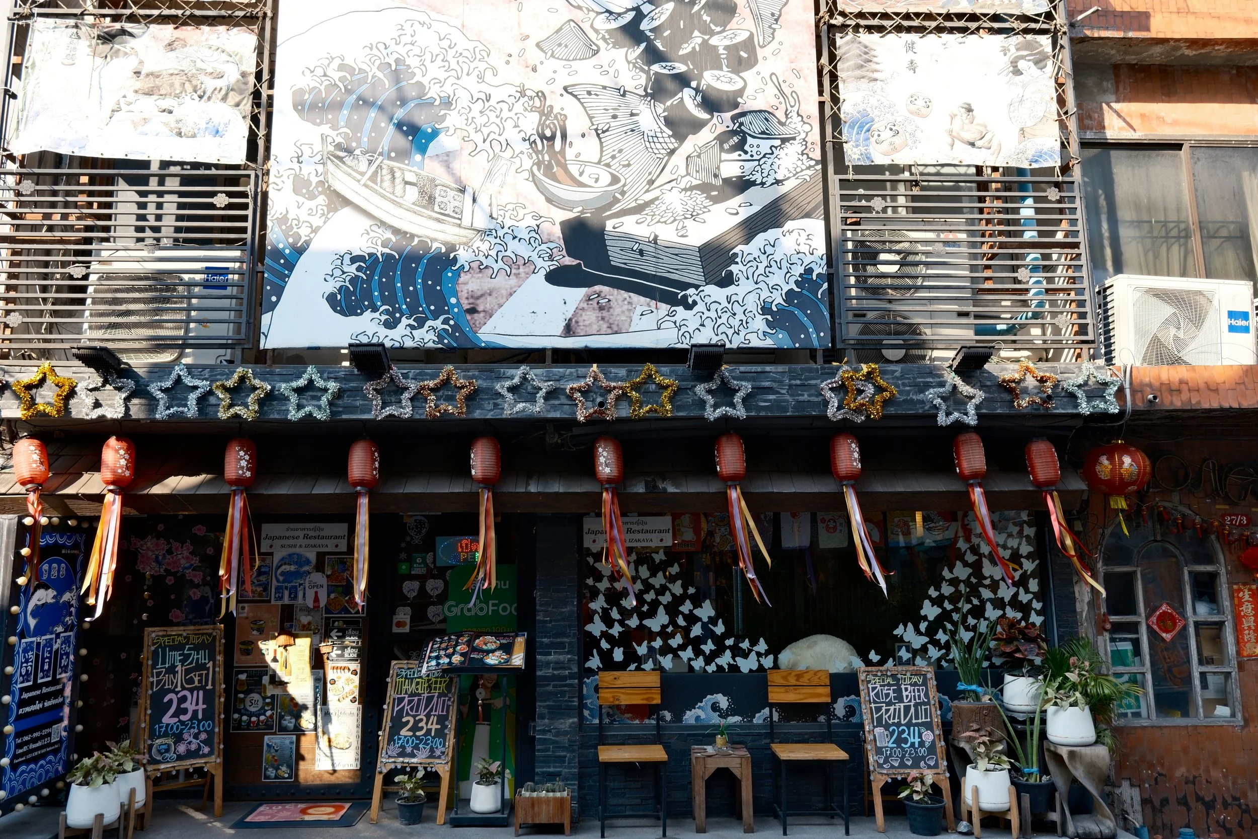 Facade of a building decorated for a celebration with red lanterns, festive star garlands, and potted plants. A large mural of a wave, boat, and book is painted on the upper level.