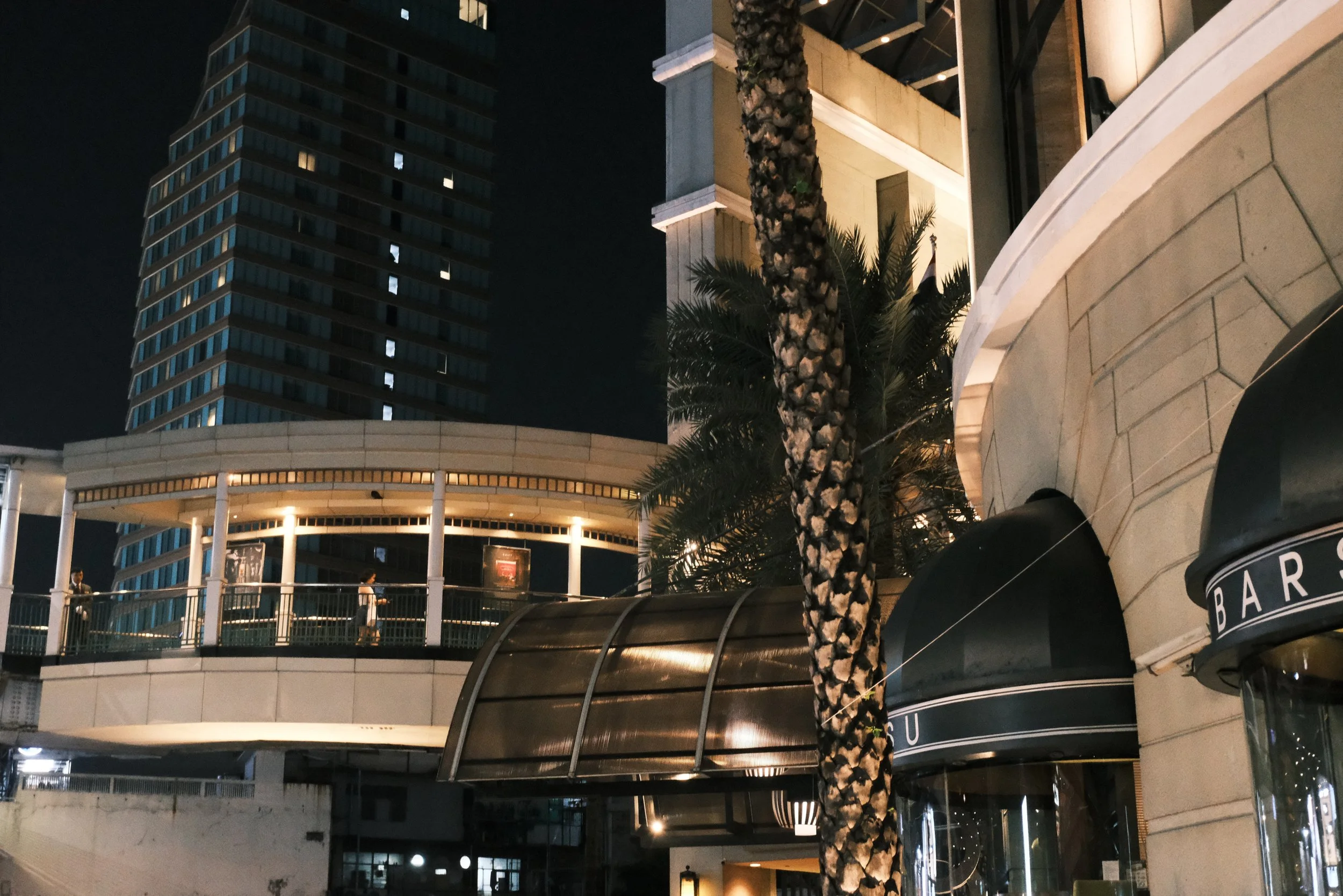 Nighttime city scene showing an elevated walkway with people, a tall building with many windows, a palm tree, and a building with a curved awning labeled 'BAR'.