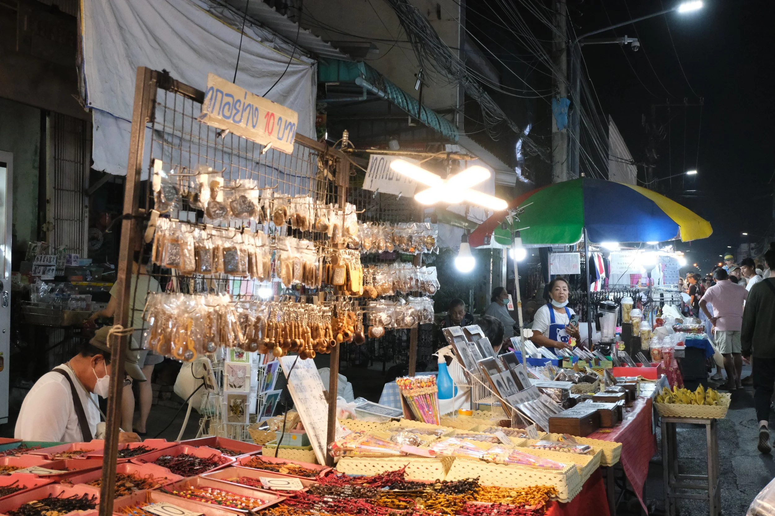 Night market scene with stalls selling jewelry, accessories, and souvenirs; colorful umbrellas and people browsing.