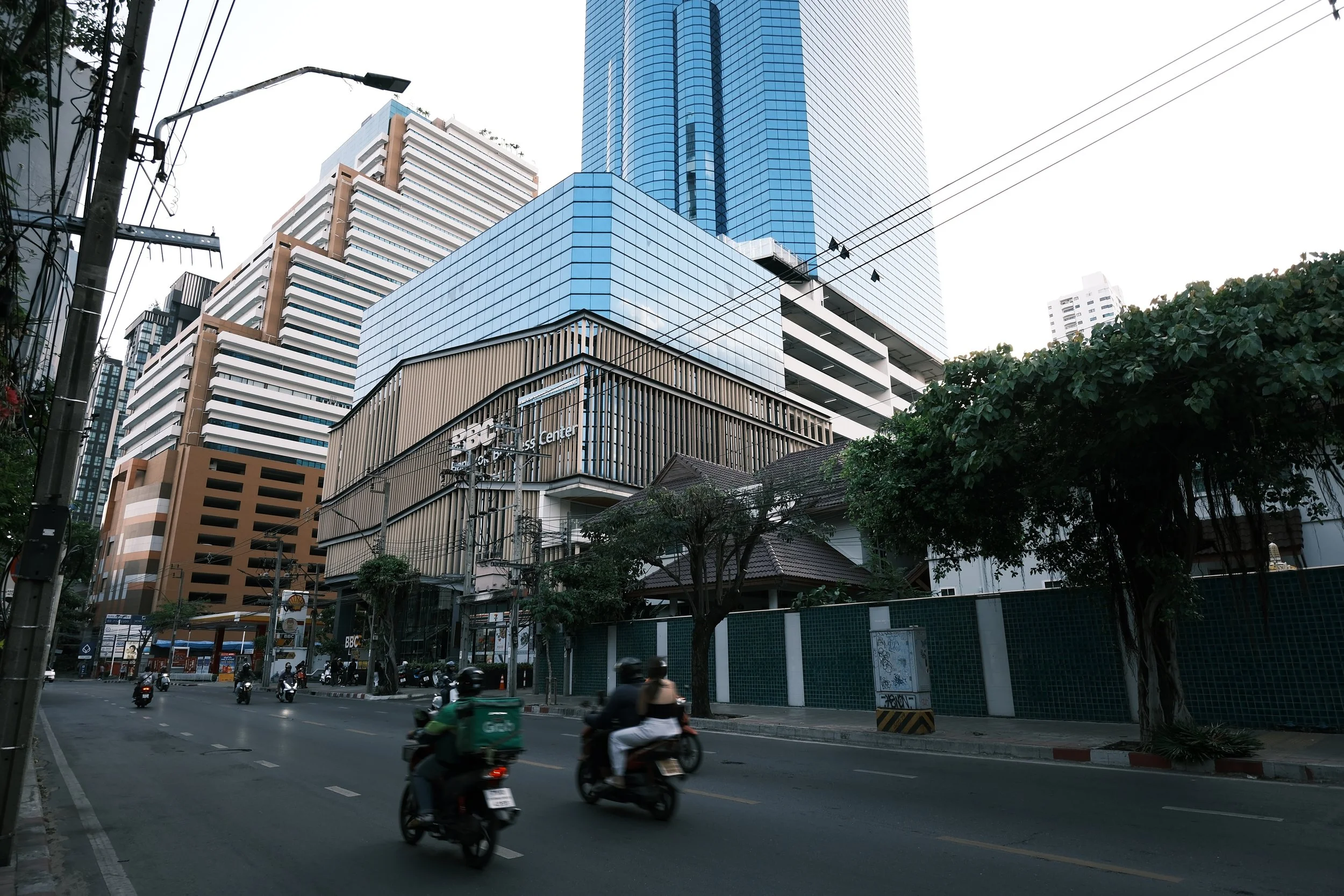 City street with high-rise buildings, some under construction, with motorcycles and cars driving along the road. Trees line the sidewalk.