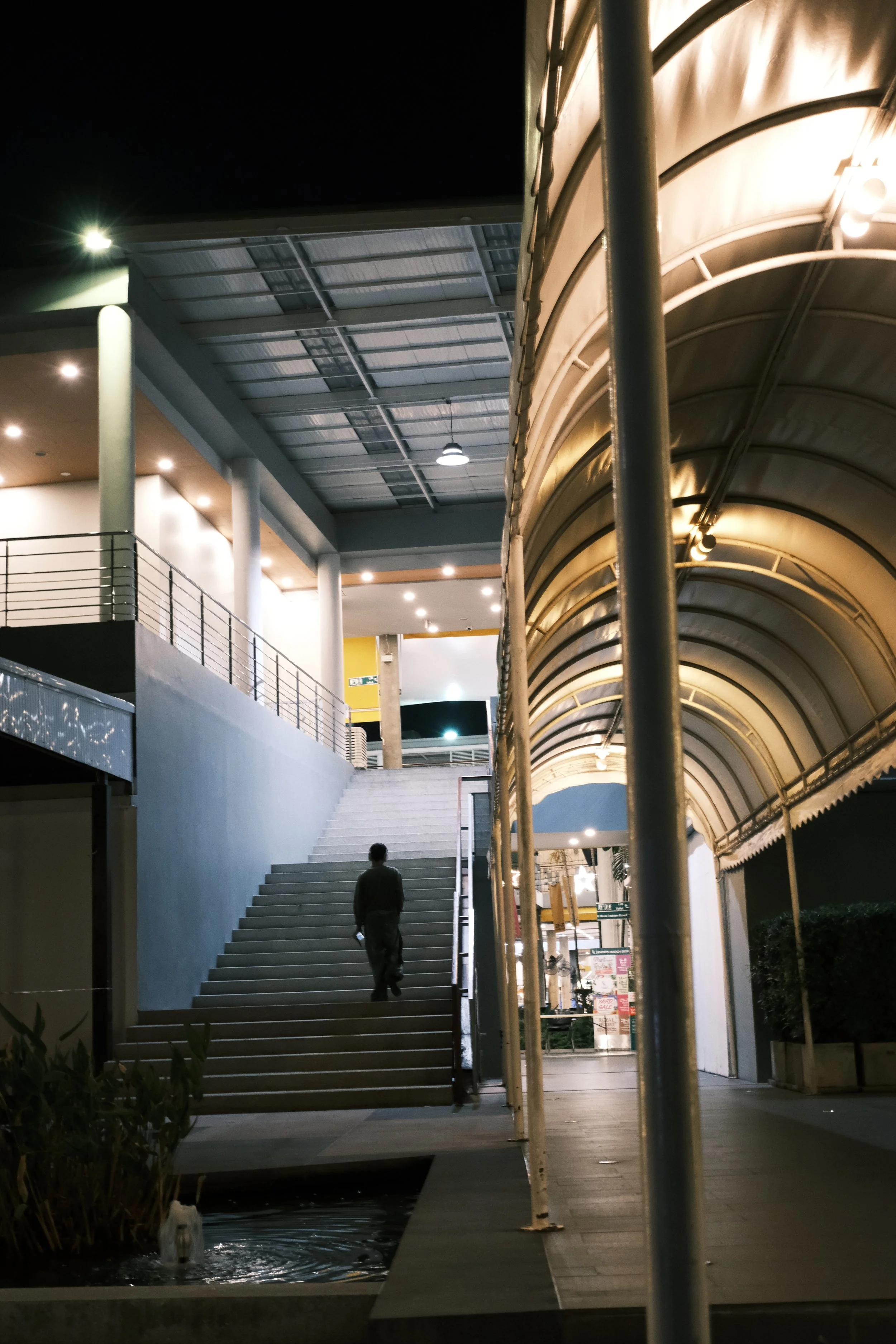 Nighttime scene of an outdoor shopping or entertainment complex with a man walking up stairs, illuminated by shop and track lighting, and an arched canopy on the right side.