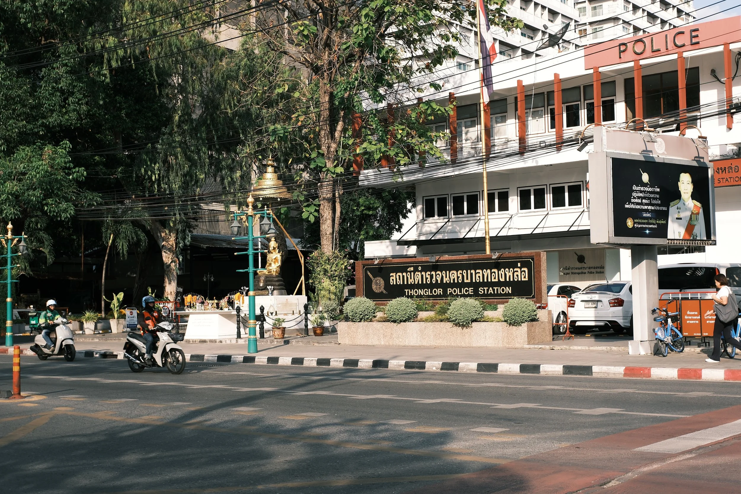 Entrance to Thonglor Police Station with a black sign and golden lettering, surrounded by greenery and street activity including motorcycles and pedestrians.