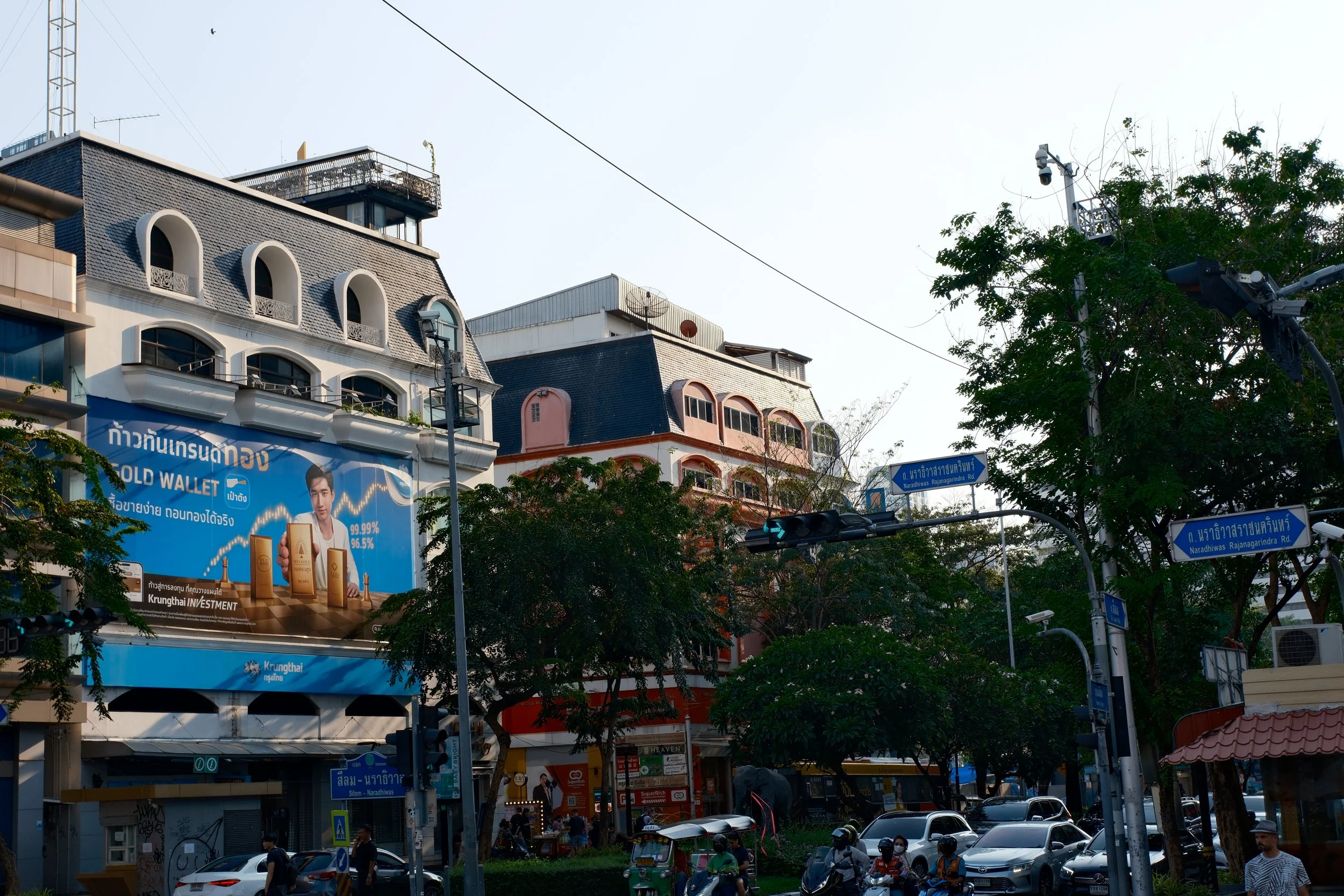 City street scene with multi-story buildings, cars, motorcycles, traffic lights, and street signs. There's a large blue billboard featuring a man and text in Thai. Trees line the street.