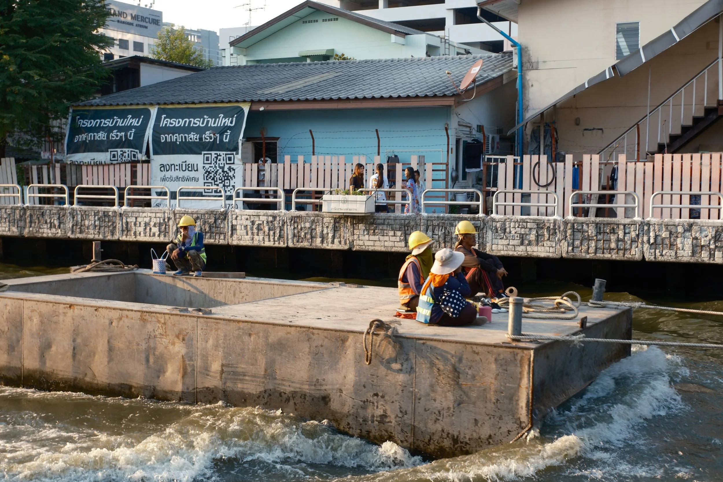 People sitting on a concrete dock by the water, wearing safety helmets and orange vests, with a building and signs in a foreign language in the background.