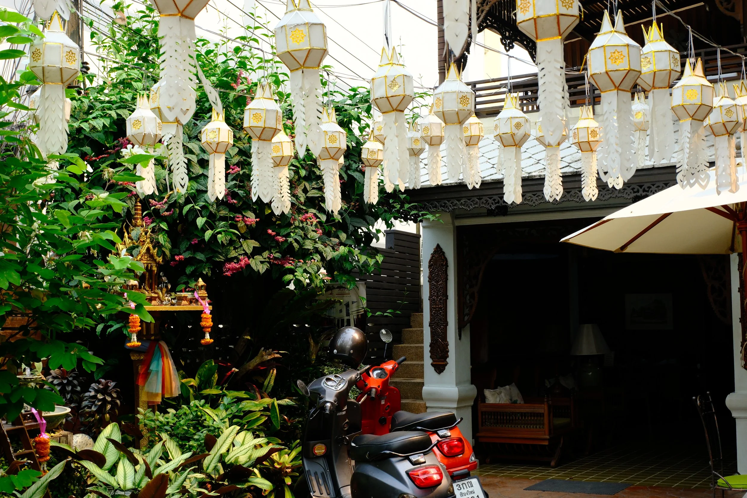 Outdoor space decorated with hanging lanterns, plants, and a scooter parked near a traditional building with ornate woodwork.