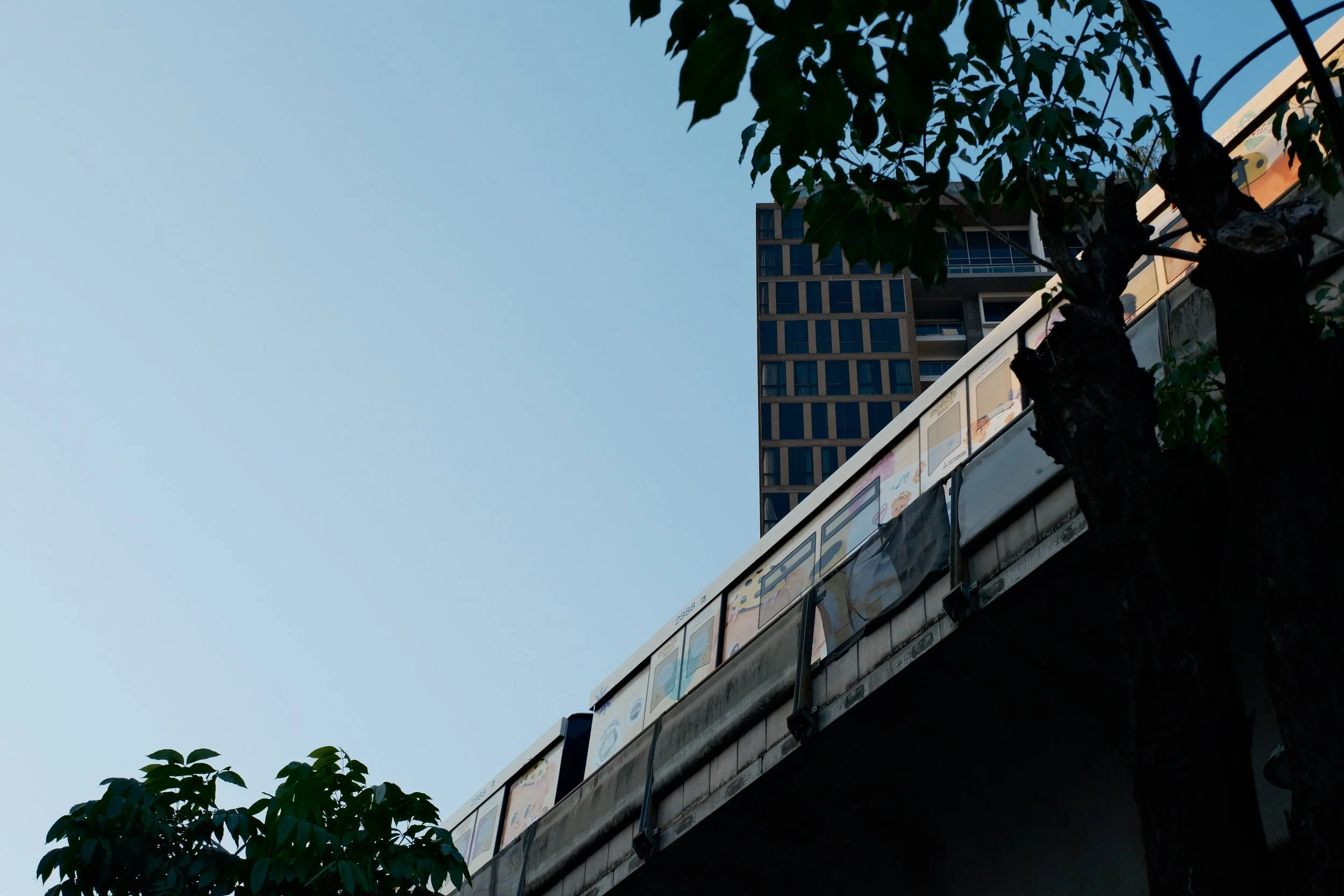 A city train traveling on an elevated track with trees in foreground and a tall modern building in the background.