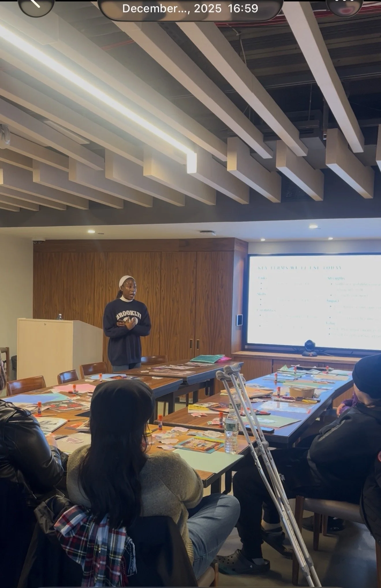 A woman stands at the front of a meeting room giving a presentation to seated attendees. The room has a large digital screen, a wooden wall, and multiple tables with paper materials, water bottles, and glue sticks.