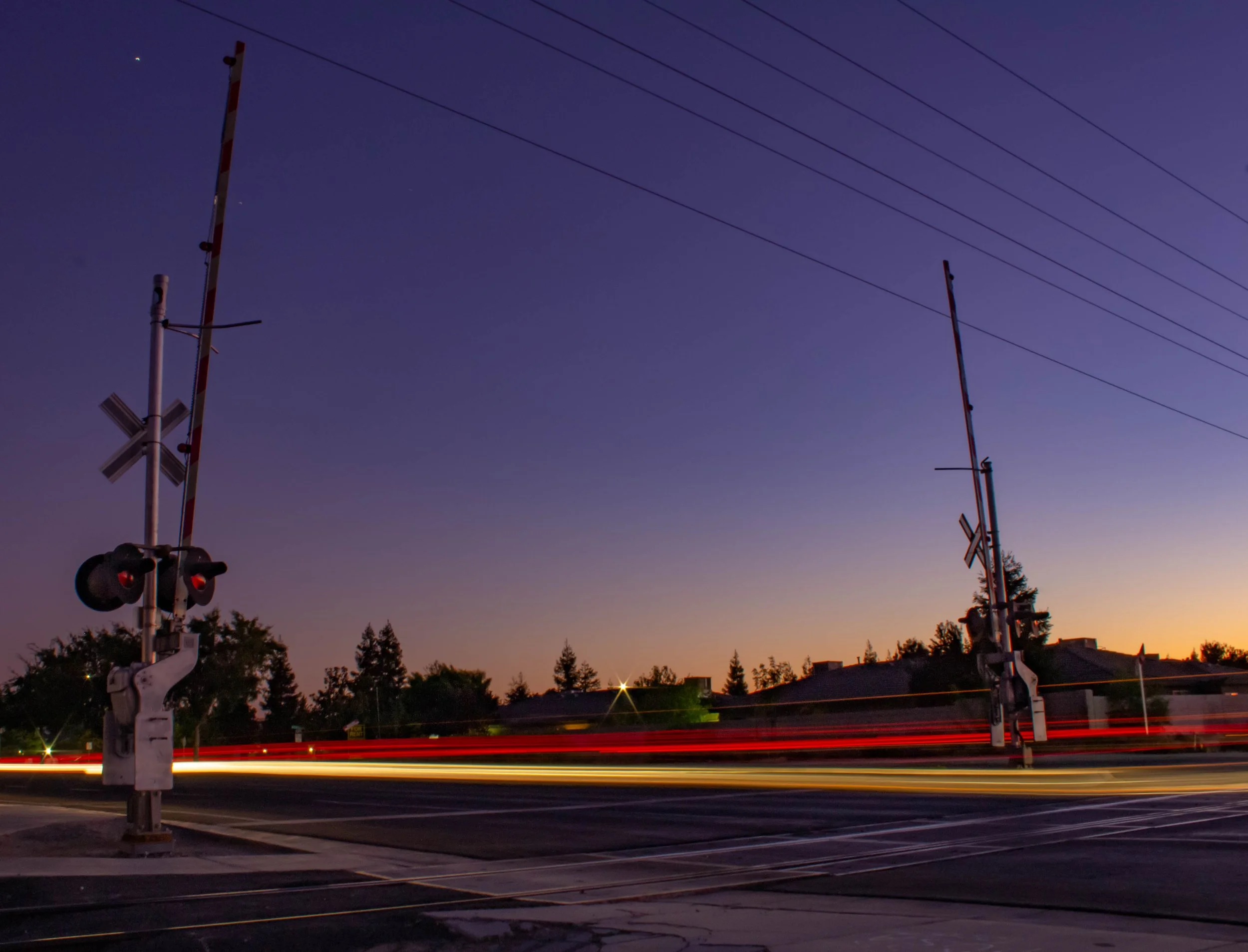 A railroad crossing at dusk with glowing red and yellow light streaks from passing vehicles, two crossing gates, and silhouetted trees and rooftops against a purple sky.