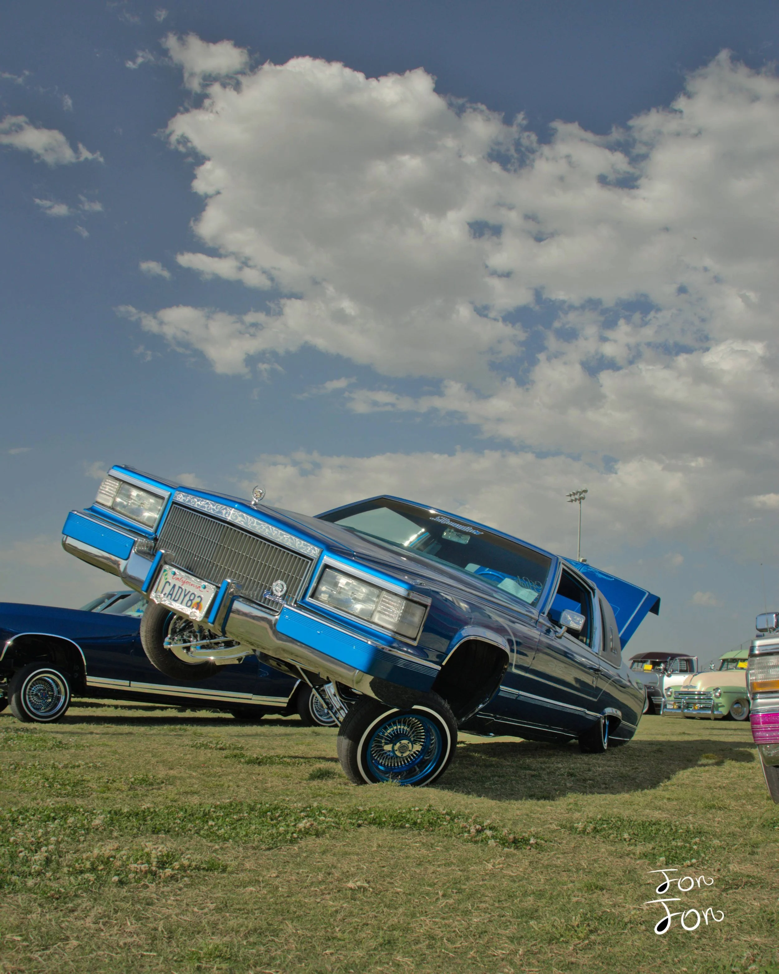 A blue, vintage car with a hydraulic lift standing on its front wheels at an outdoor car show with other classic cars parked in the background and a partly cloudy sky above.