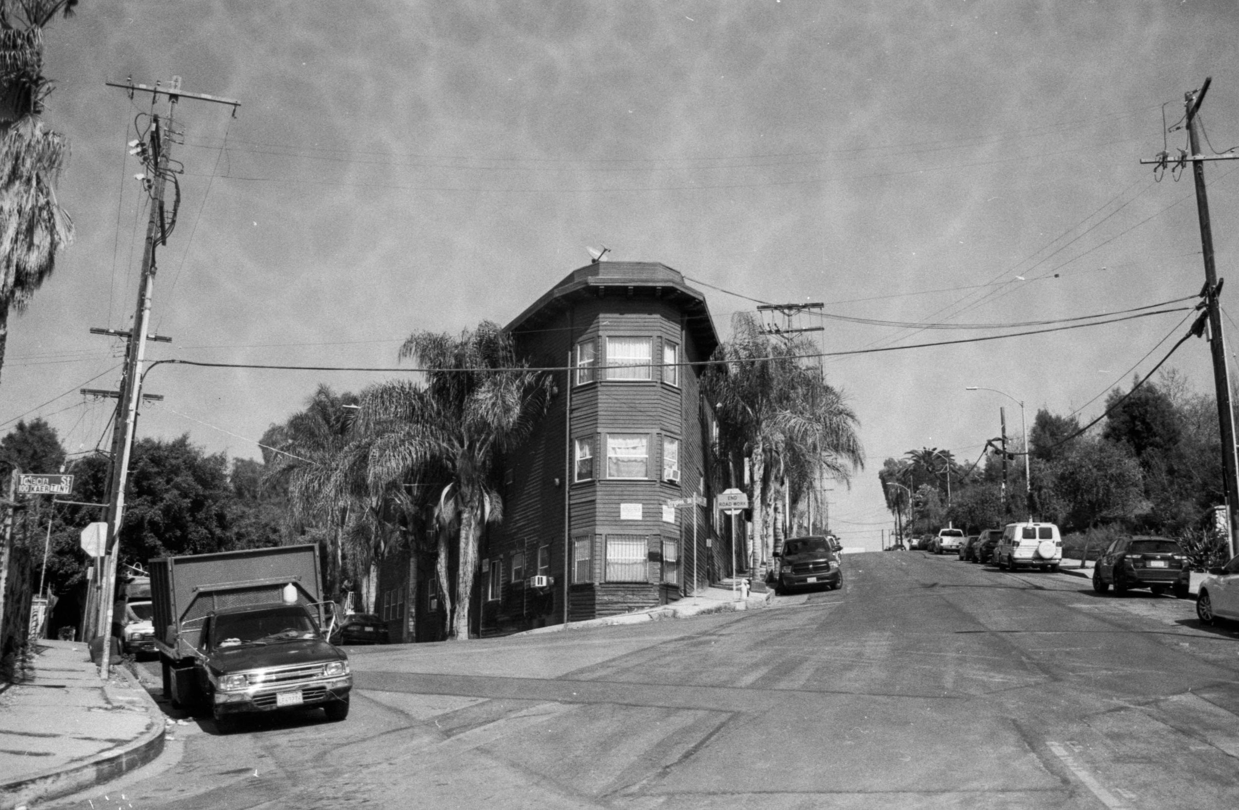 Black and white photograph of a residential street corner with a tall, narrow wooden building, several parked cars, utility poles and trees, and a clear sky.