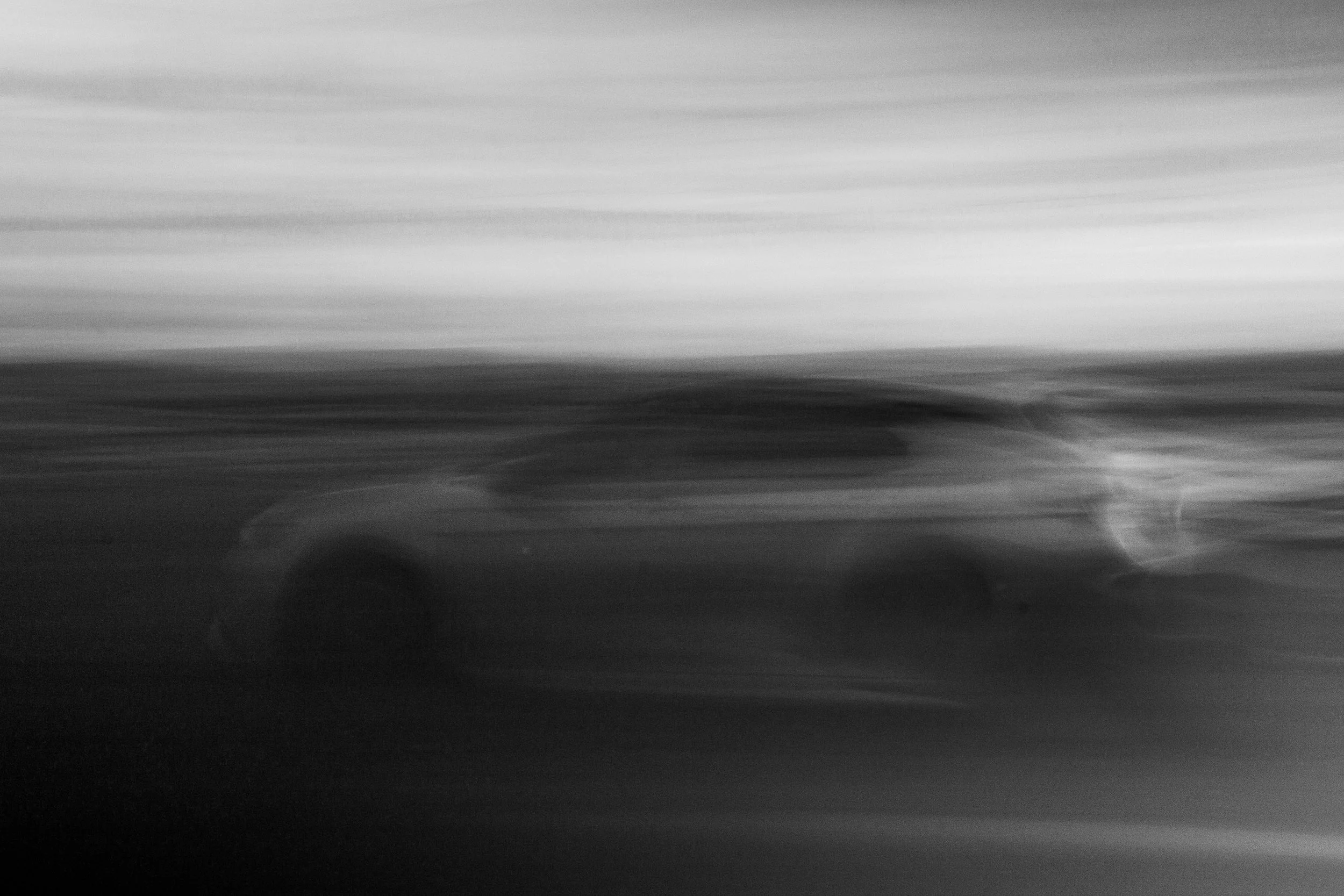 A blurred black-and-white photo of a car in motion with streaks of clouds in the sky background.