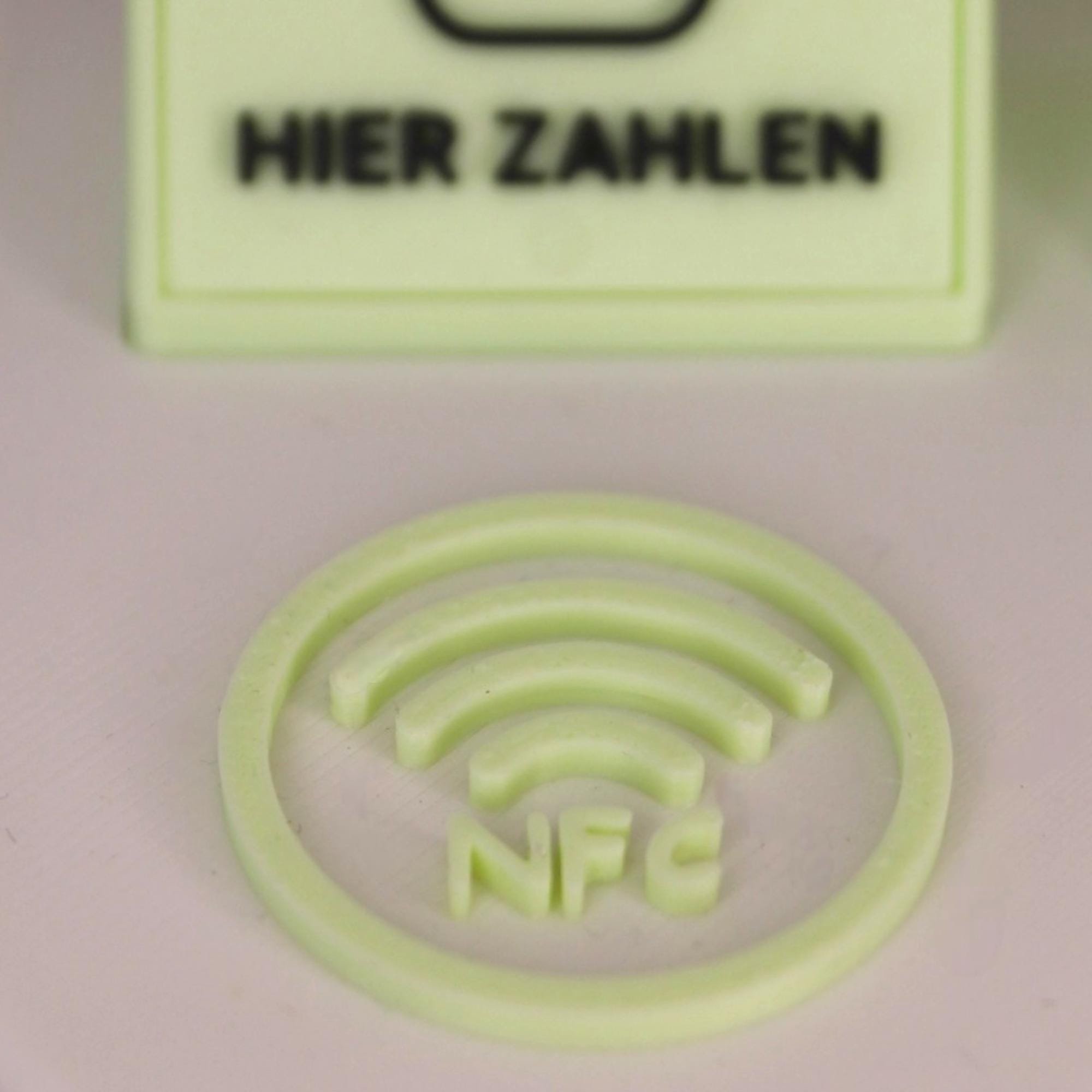 Nahaufnahme des Inlays Hier zahlen und der runden NFC-Fläche am pastellgrünen Aufsteller auf neutralem Hintergrund.