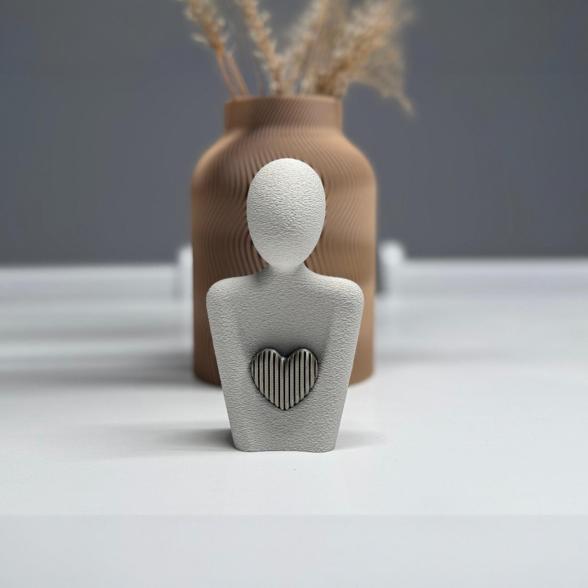 Weiße Herzfigur mit grauem Herzdetail auf gedecktem Tisch vor Vase mit Trockenblumen, dekorativ inszeniert für modernes Wohnen.