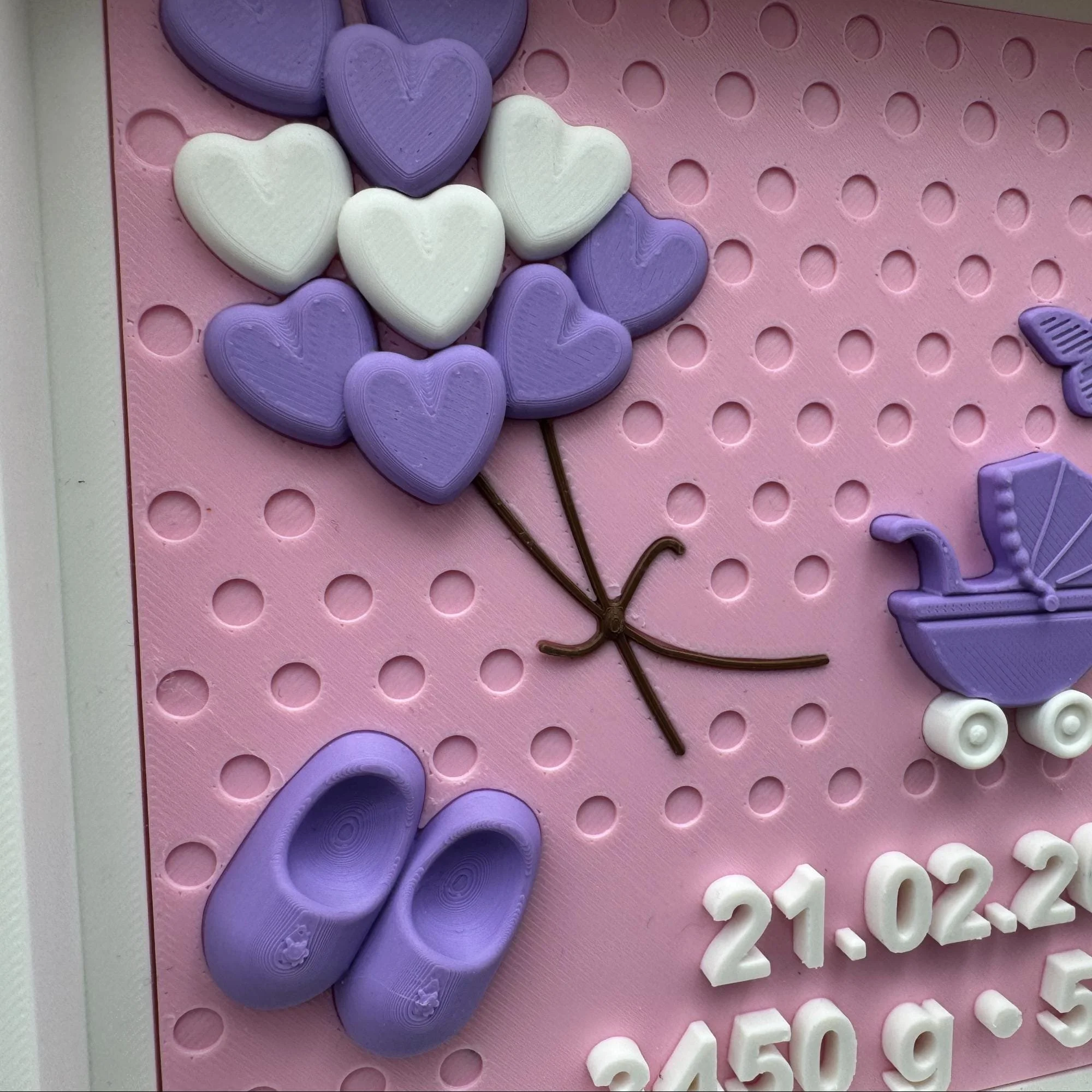 Detail der Geburtstafel mit lilafarbenen Herzballons, kleinem Kinderwagen und geprägten Geburtsdaten auf rosa Hintergrund.