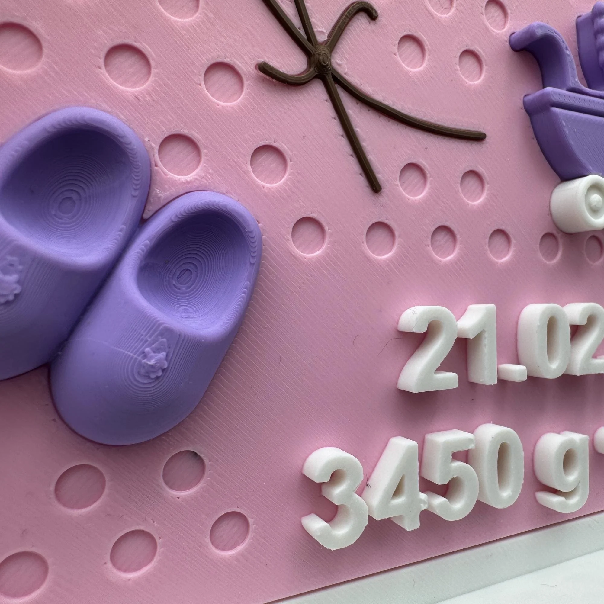 Nahaufnahme mit lilafarbenen Babyschuhen, Teil des Kinderwagenmotivs und erhabenen Geburtsdaten auf strukturierter rosa Fläche.