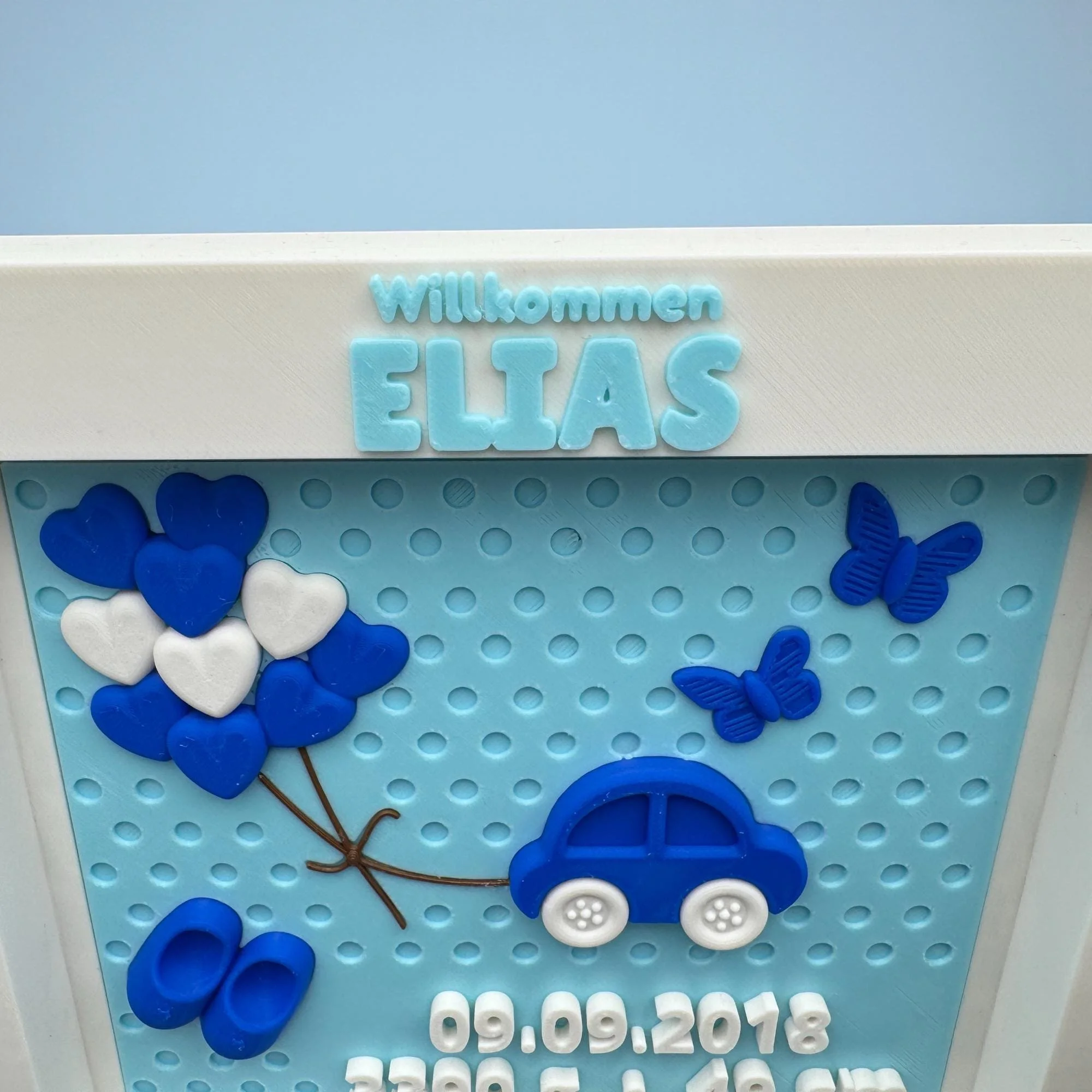 Detail der personalisierten Geburtstafel mit erhabenem Namen Elias, blauen Wolkenballons, Auto und Schmetterlingen auf hellblauem Hintergrund