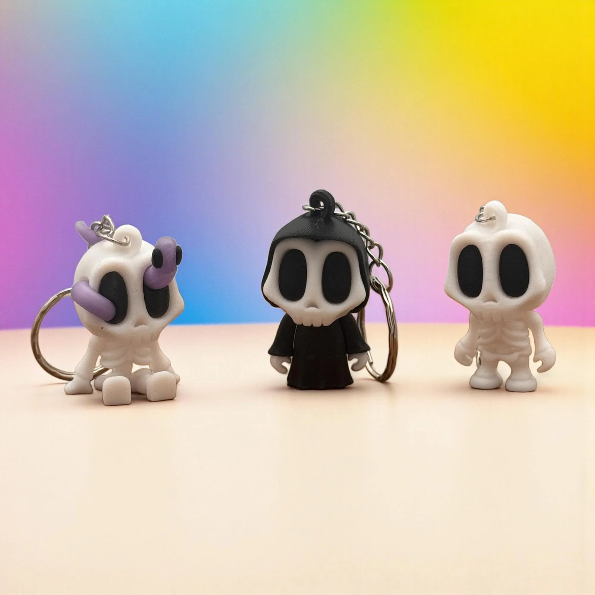 Drei Skelett Schlüsselanhänger nebeneinander auf hellem Untergrund vor buntem Farbverlauf: links sitzende Figur mit lila Wurm, in der Mitte schwarze Figur, rechts weißes Skelett.