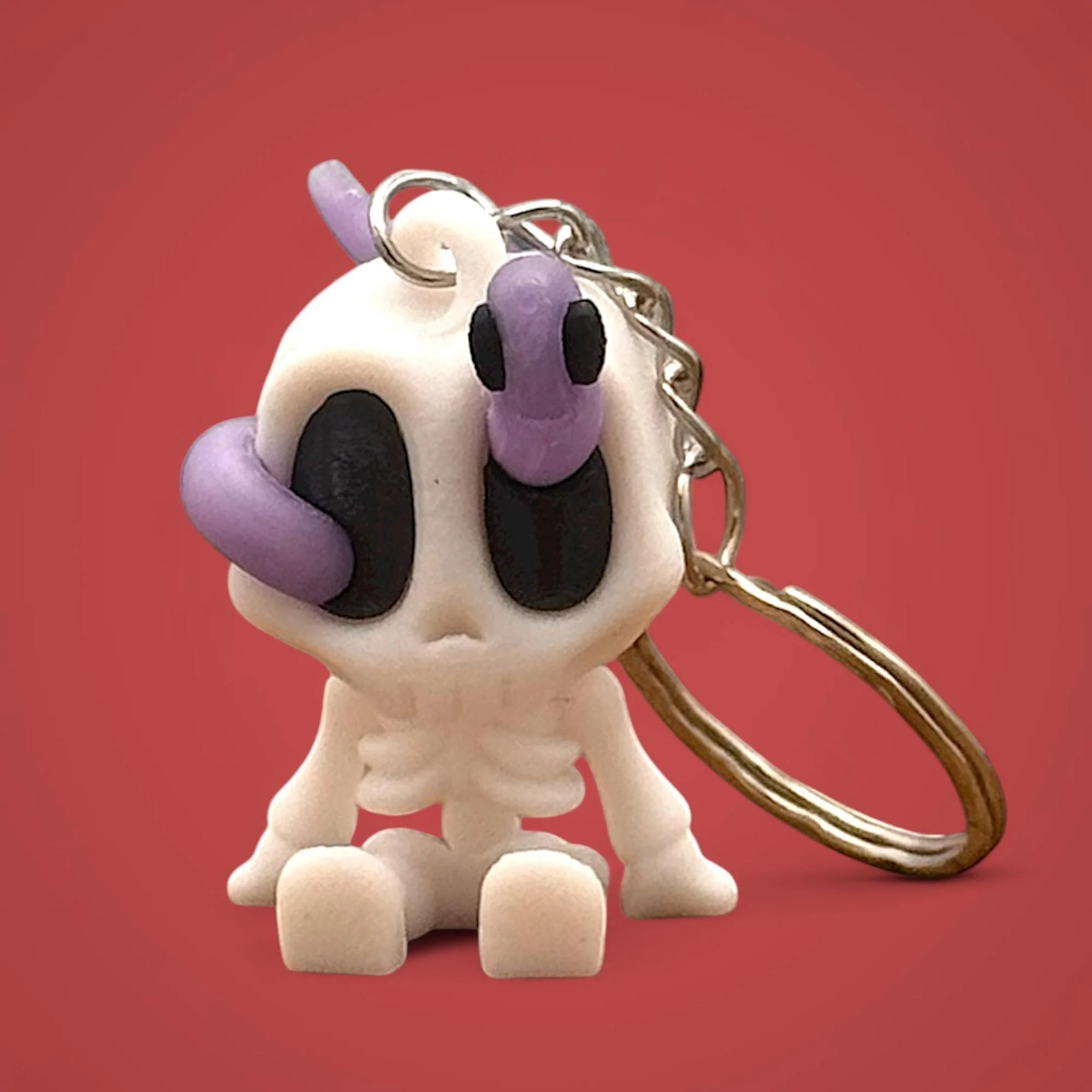 Sitzender Skelett Schlüsselanhänger mit lila Wurm auf dem Kopf und Metallring vor rotem Hintergrund.