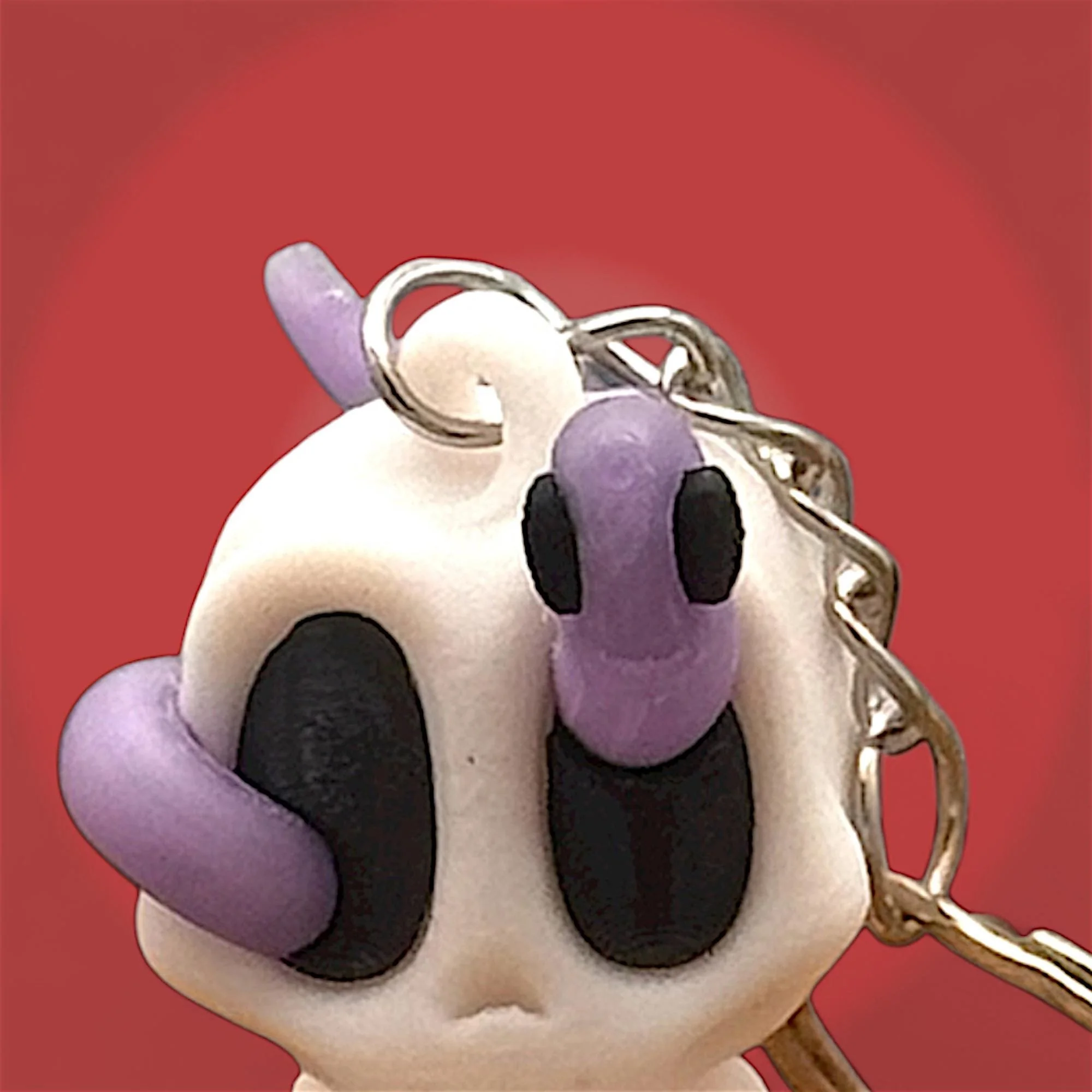Nahaufnahme eines sitzenden Skelett Schlüsselanhängers mit lila Wurm auf dem Kopf und Metallring vor rotem Hintergrund.