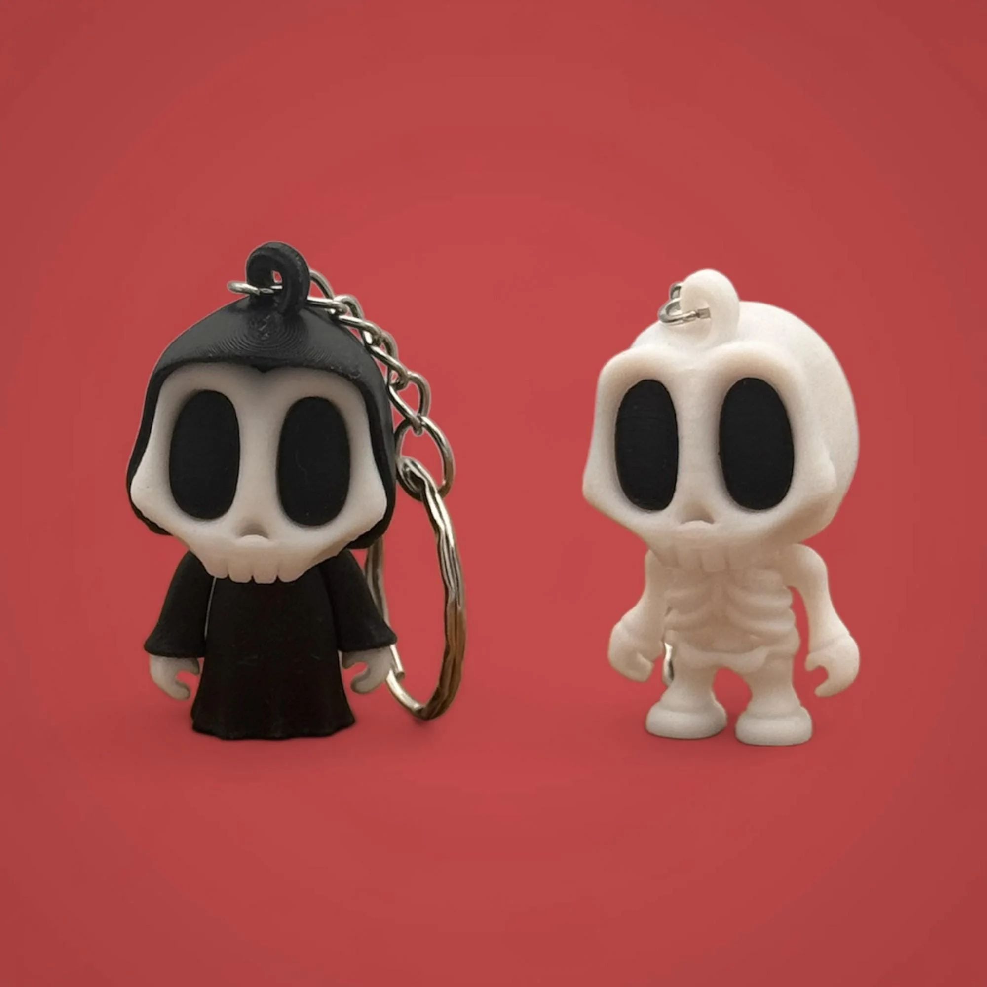 Zwei Skelett Schlüsselanhänger nebeneinander vor rotem Hintergrund: links schwarze Figur, rechts weißes Skelett.