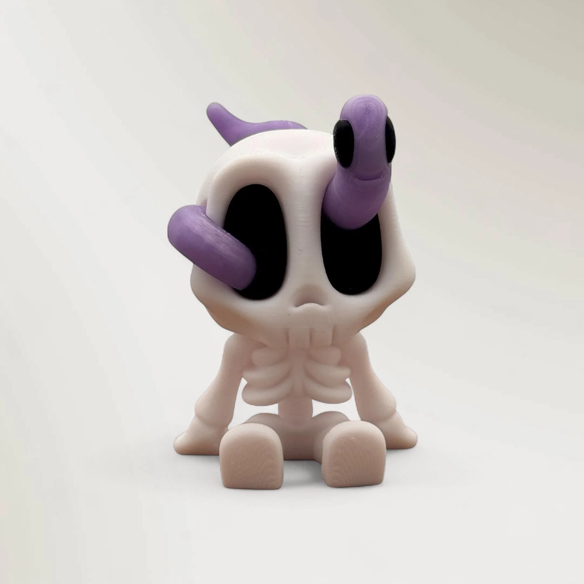 Sitzendes weißes Mini-Skelett mit schwarzer Augenpartie und lila Wurm auf dem Kopf vor hellem Hintergrund. Die Figur ist leicht seitlich gedreht und vollständig sichtbar.