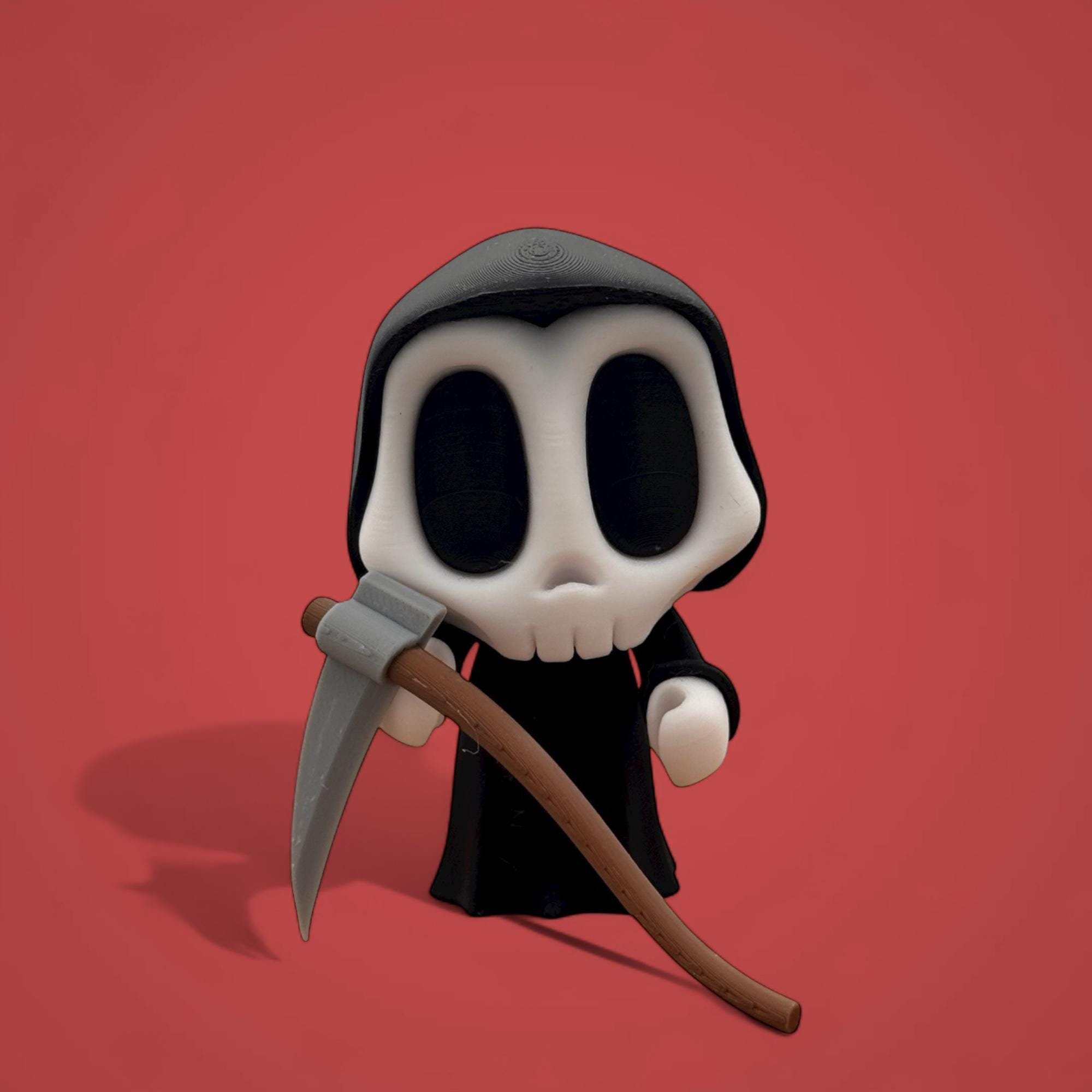 Schwarzes Mini-Skelett mit heller Gesichtsfläche und großer Sense steht vor rotem Hintergrund. Die Figur ist frontal fotografiert und hält die Sense schräg vor dem Körper.