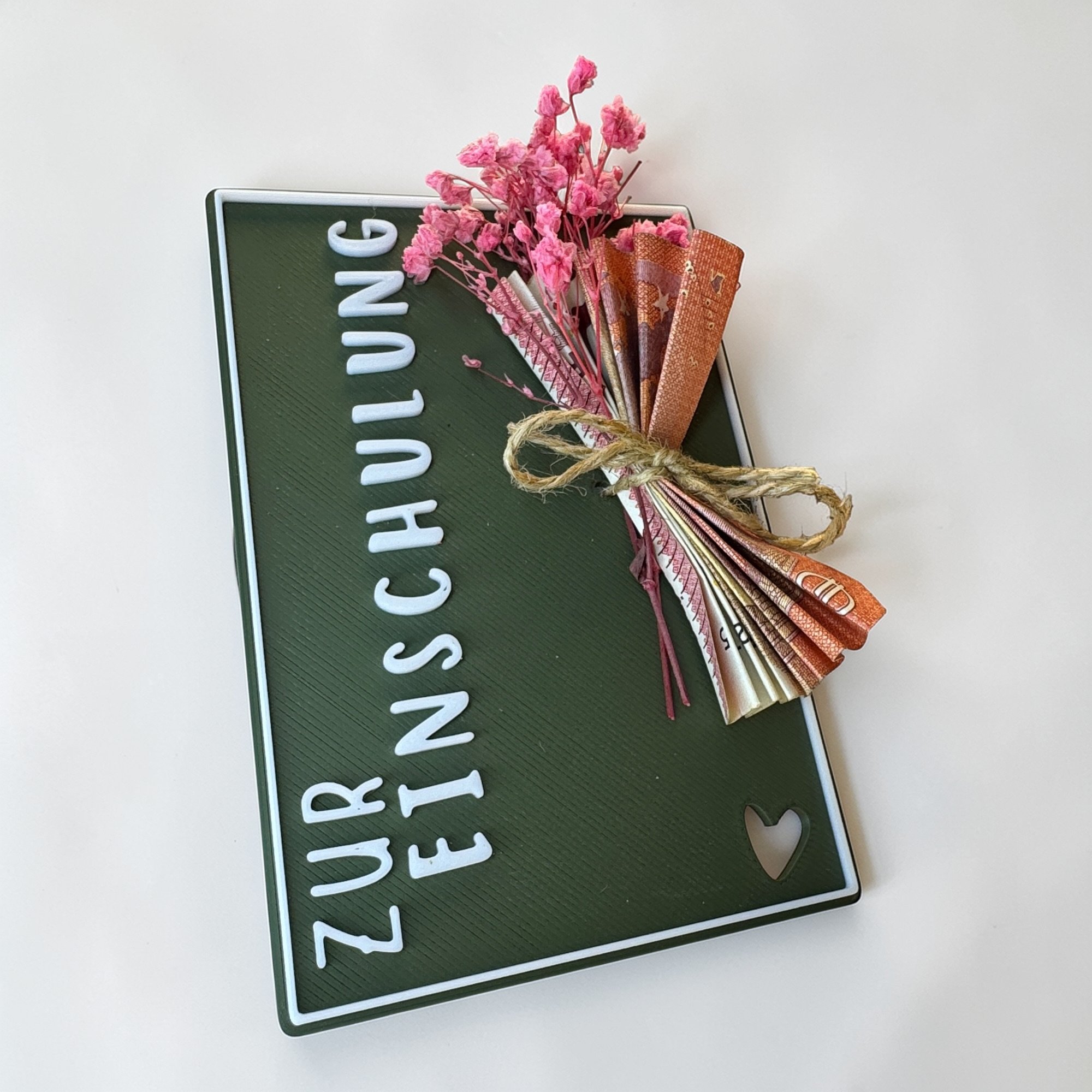 Dunkelgrüne Geschenkkarte zur Einschulung mit hochkantem Schriftzug, Kordelschleife und rosa Blütenstrauß auf hellem Hintergrund.