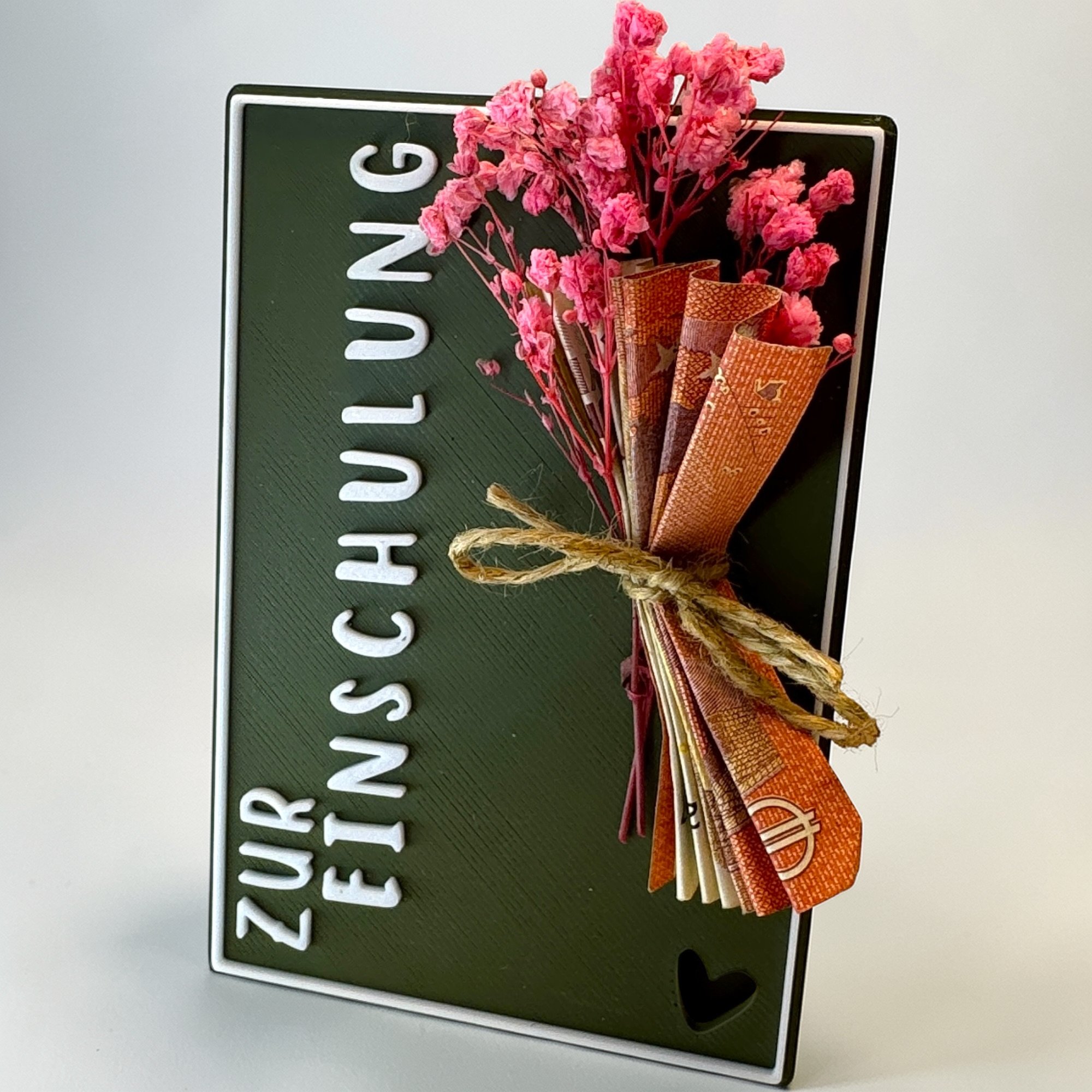 Schräg liegende dunkelgrüne Karte Zur Einschulung mit weißem Schriftzug, Kordel und rosa Blüten als dekorativem Detail.