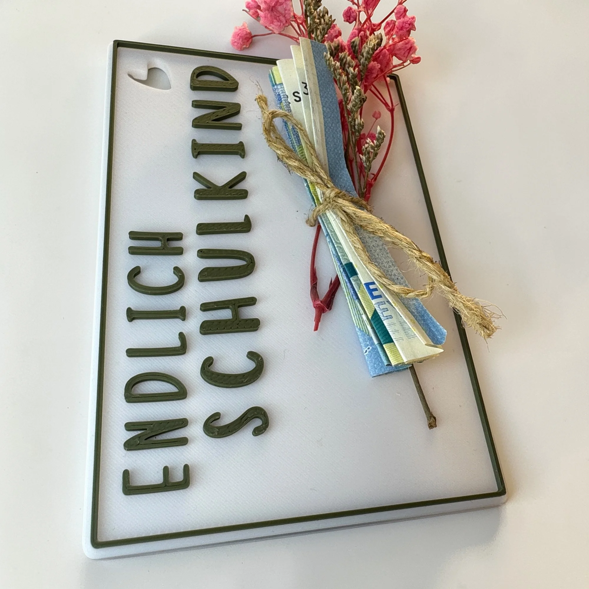 Weiße Geschenkkarte Endlich Schulkind mit senkrechter Schrift und kleinem Blütenstrauß, leicht schräg auf hellem Untergrund.