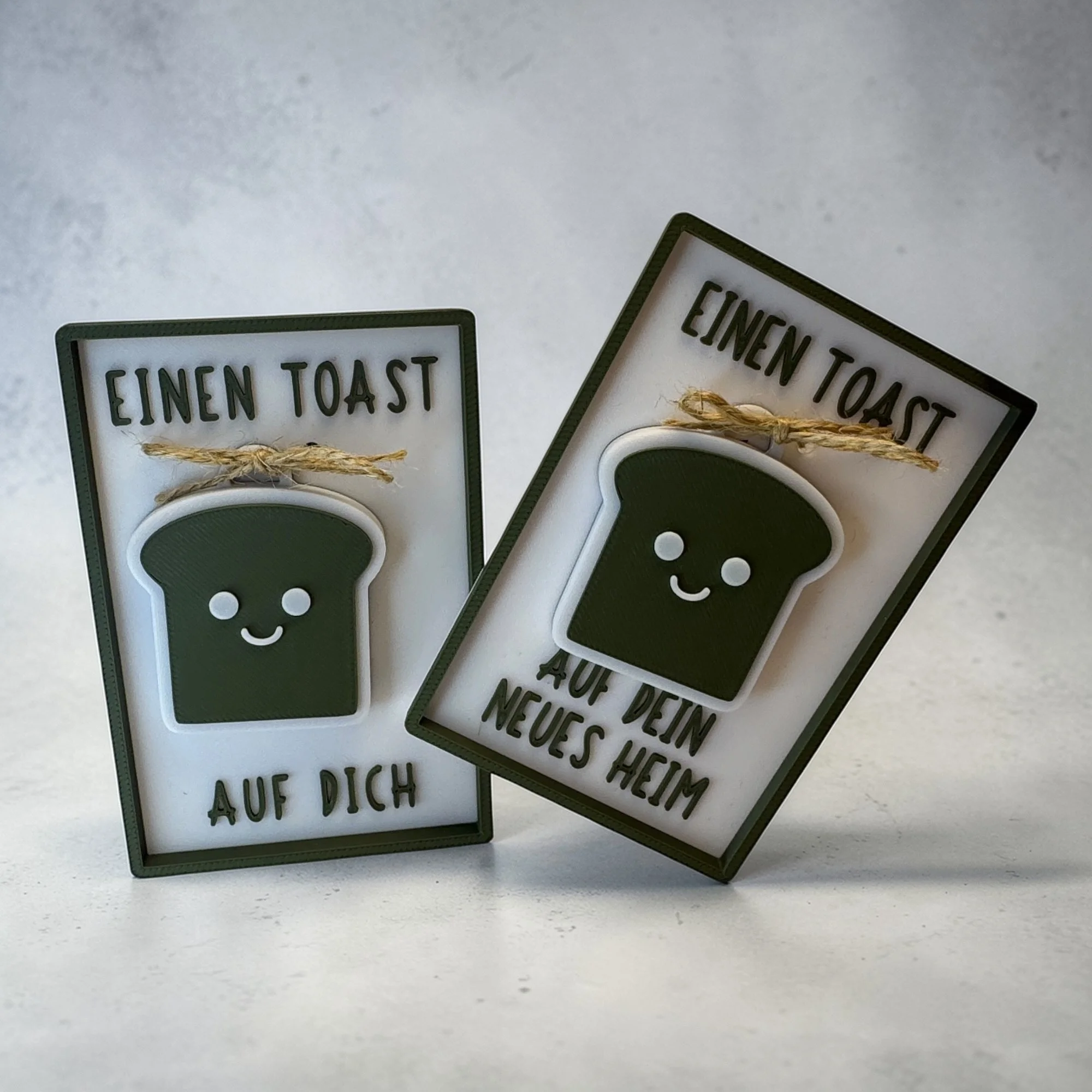 Zwei Einzugskarten mit Toast-Schlüsselanhänger nebeneinander, in Weiß und Olivgrün als originelle Glückwunschkarte für Einzug oder Umzug.