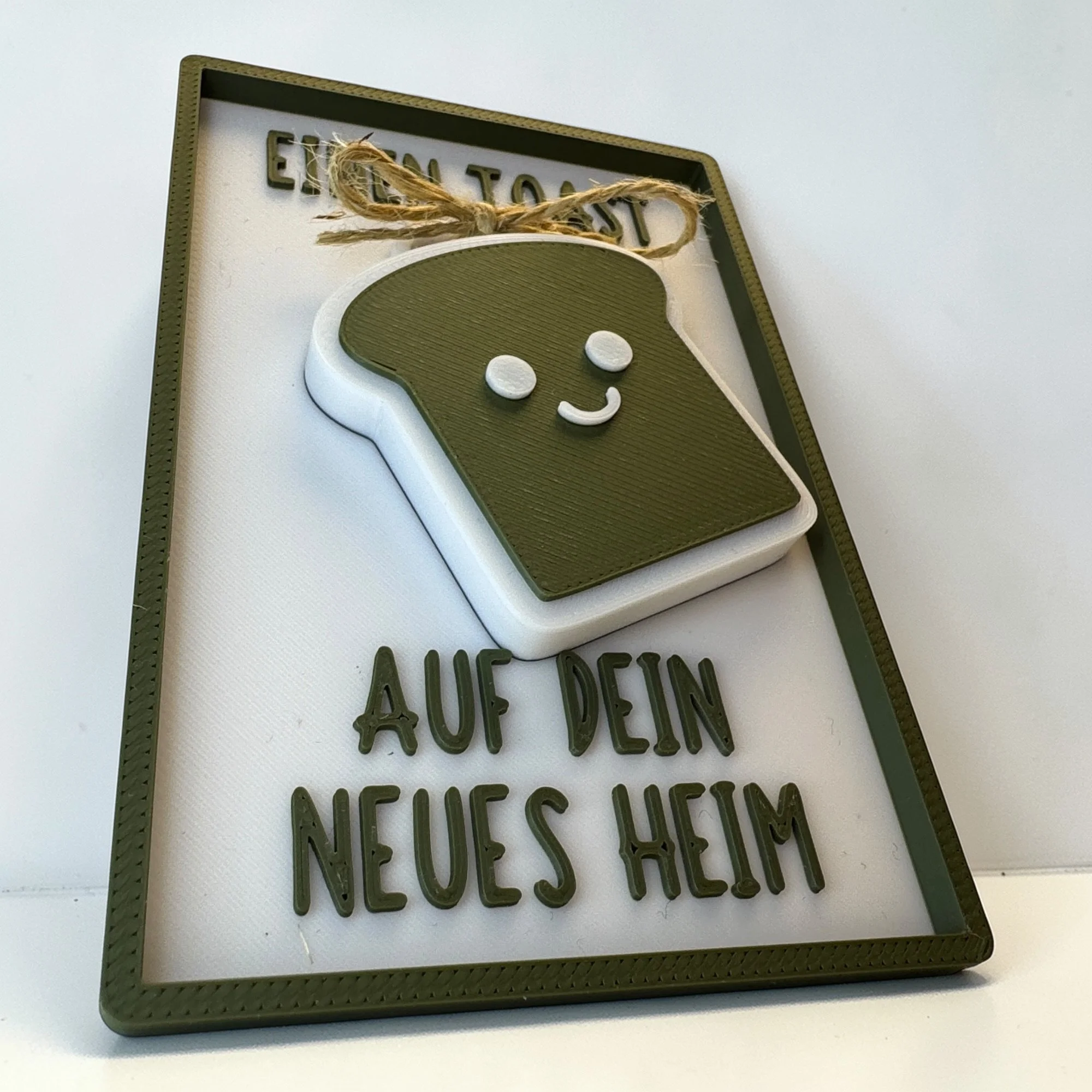 Schräg stehende Einzugskarte mit Toast-Schlüsselanhänger und Spruch Einen Toast auf dein neues Heim in Weiß und Olivgrün.