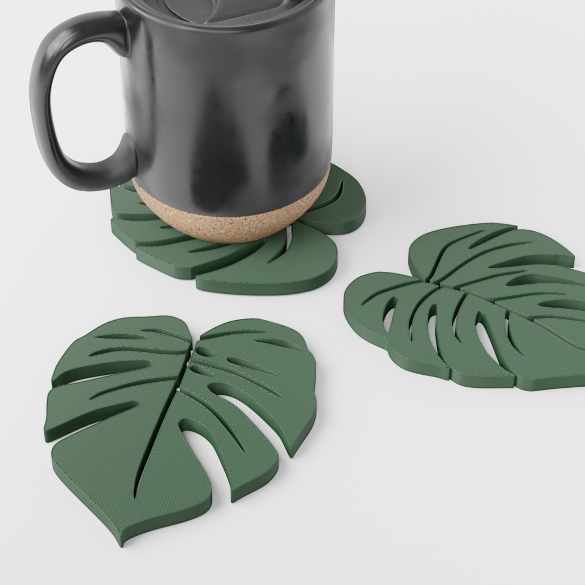 3D-gedrucktes Monstera-Untersetzer-Set mit schwarzer Tasse auf einem Blatt-Untersetzer und zwei weiteren grünen Designs auf hellem Hintergrund