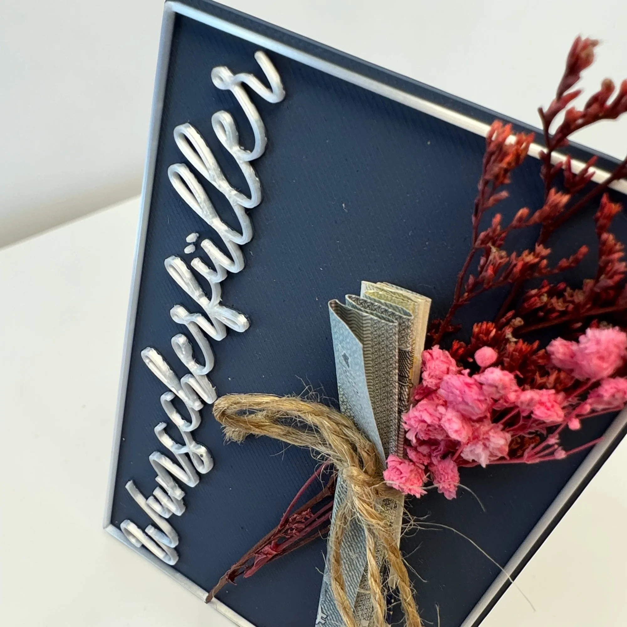 Detail der Wunscherfüller Karte mit weißem Schriftzug, Juteband, rosa Trockenblumen und befestigtem Geldschein an der rechten Seite