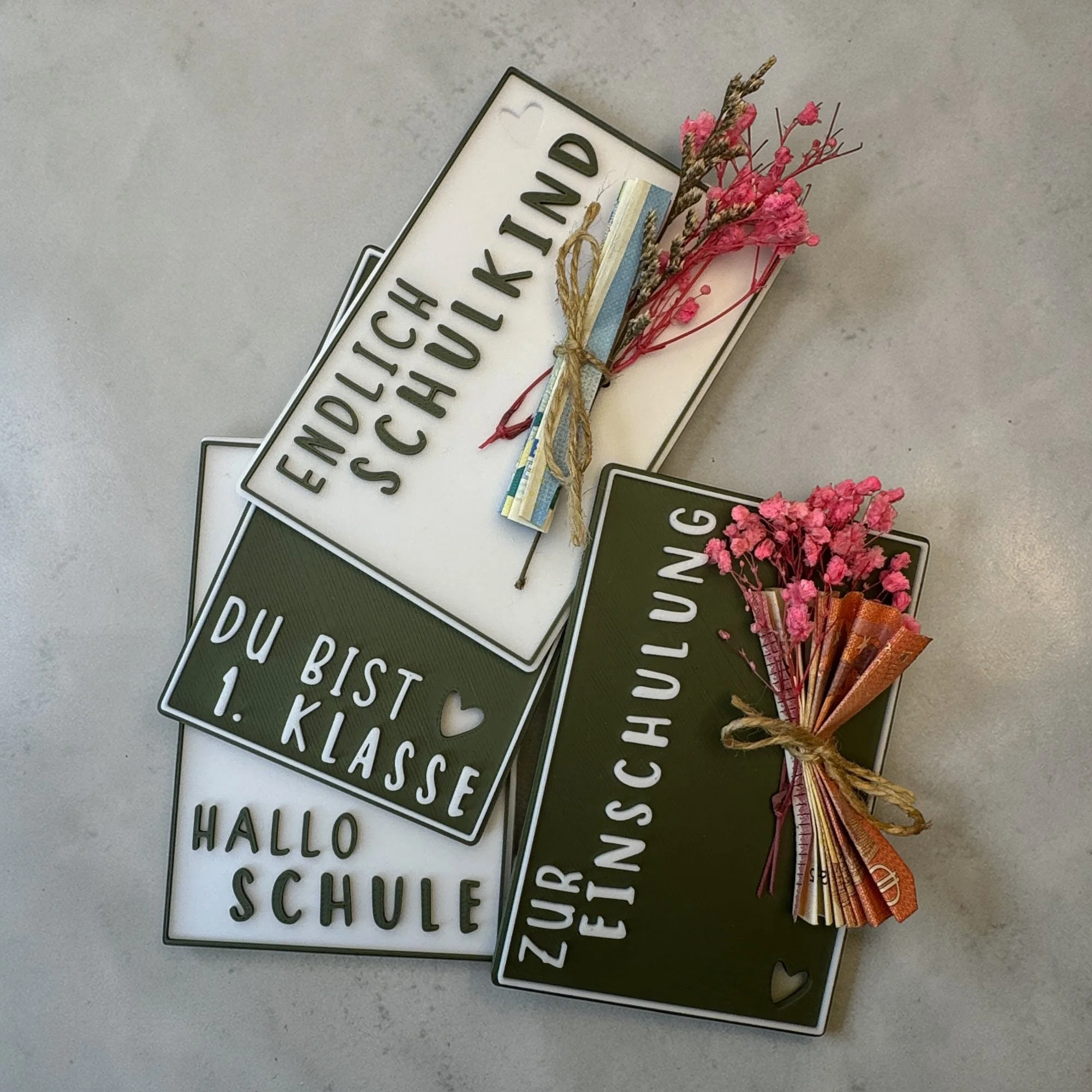 Übersicht mit mehreren Geschenkkarten zur Einschulung in Weiß und Dunkelgrün mit Blütenstrauß und unterschiedlichen Schriftzügen.
