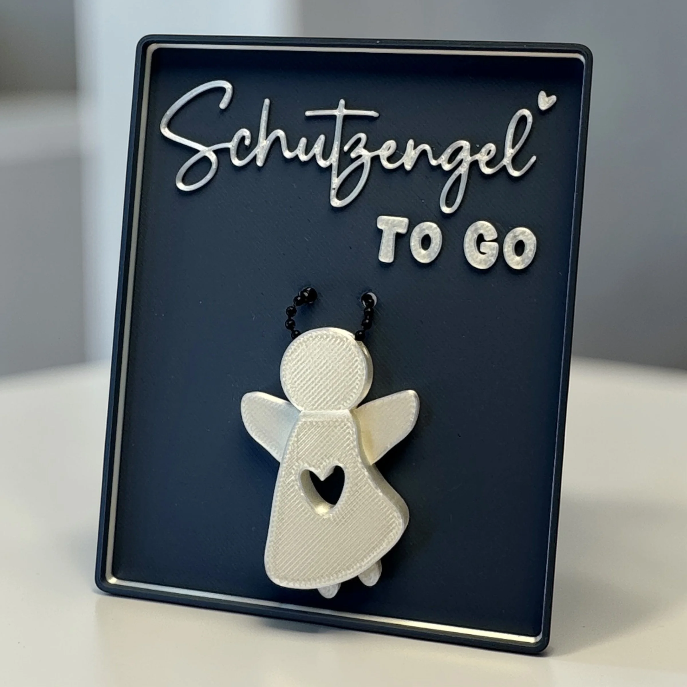 Schutzengel Anhänger in dunkelblauer Spruchschale mit Schriftzug Schutzengel to go, frontal dargestellt als kleine Geschenkidee mit Herzdetail.