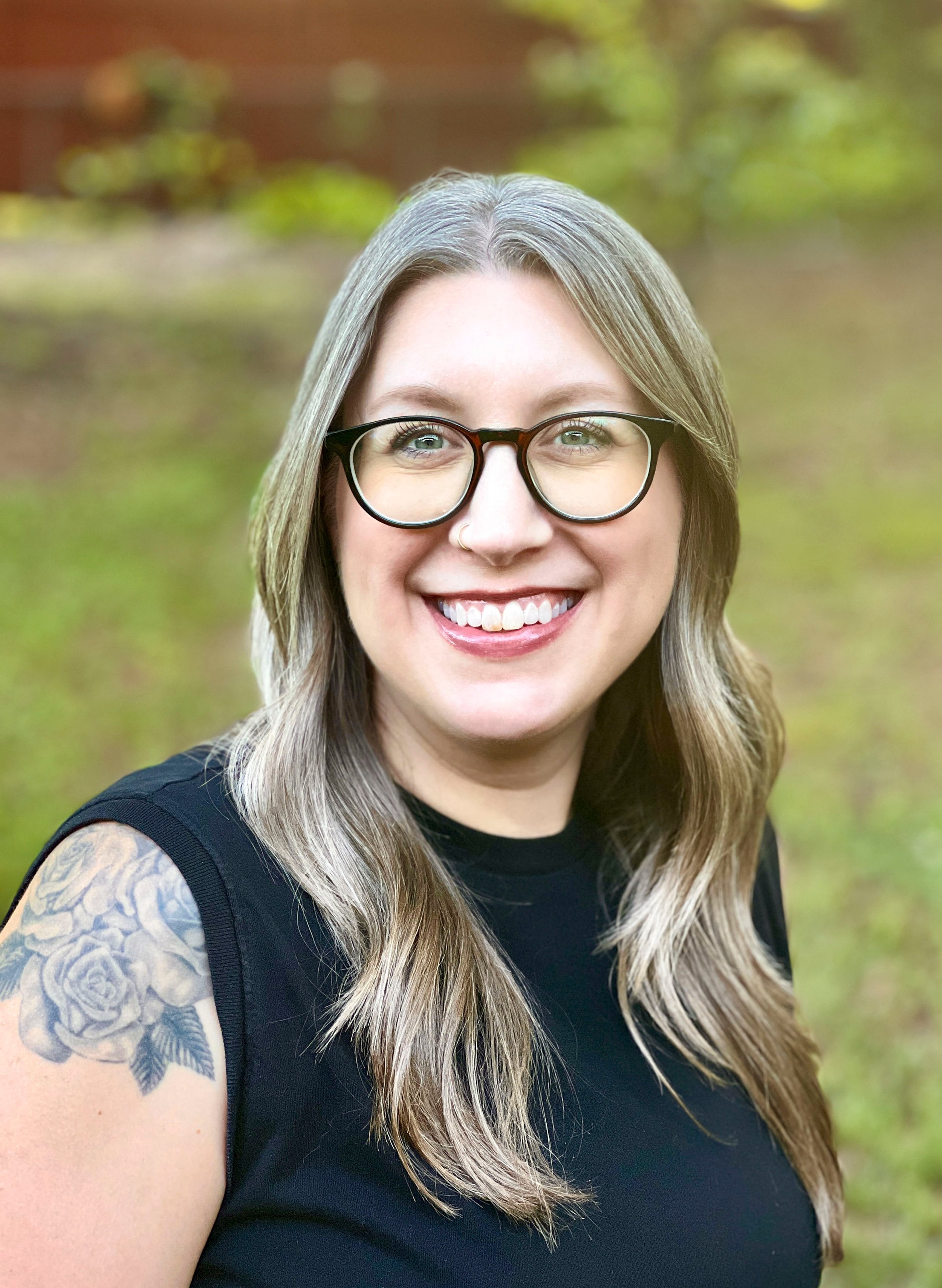 Close-up portrait of a woman with gray hair, glasses, and a nose ring, smiling outdoors with a blurred green and brown background.