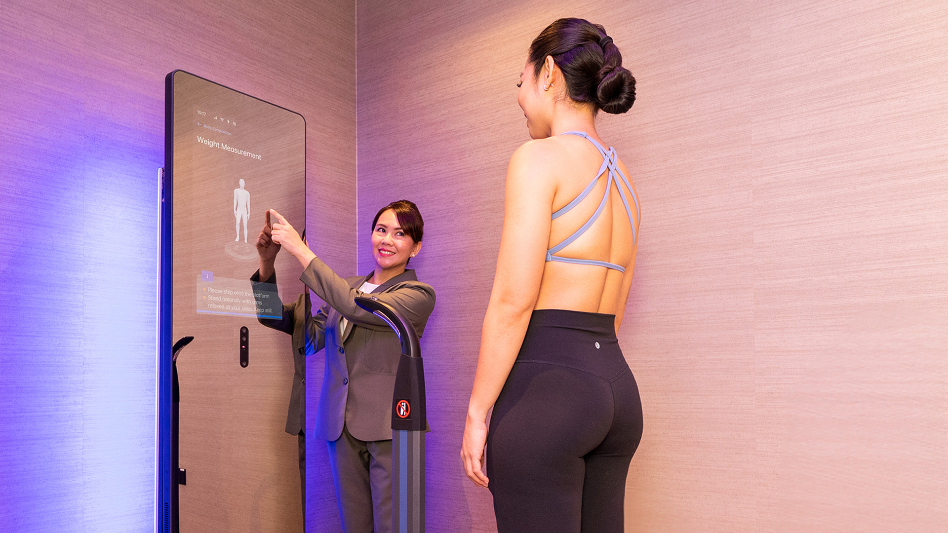 A woman in workout clothes standing on a scale while a technician uses a large digital body composition scanner to measure her weight and body data in a modern fitness facility.