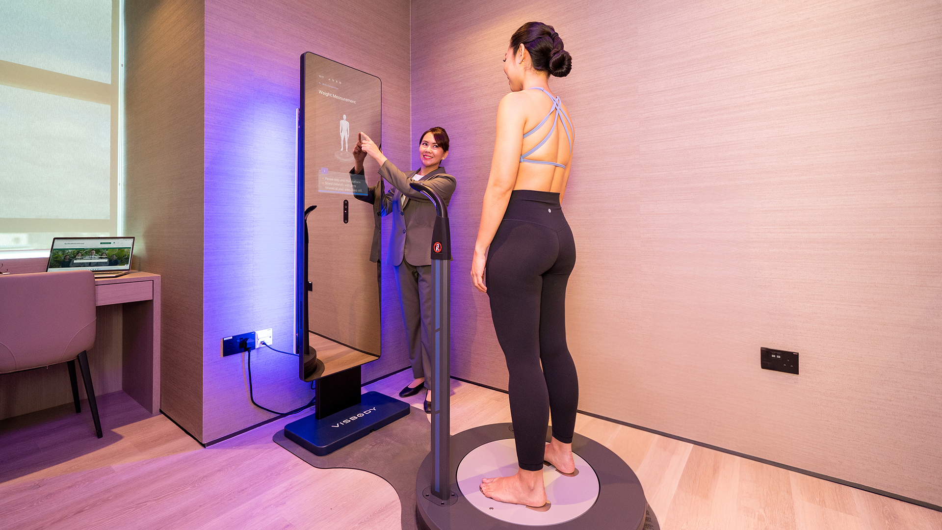 A woman in athletic wear stands on a body composition scanner as a technician in a suit uses the touchscreen to operate it in a R17 treatment room.