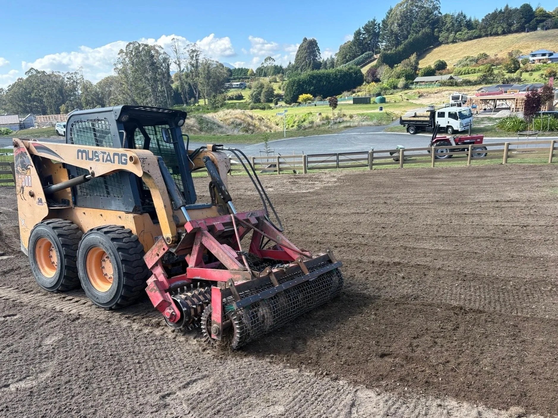 A small construction skid steer loader working on tilled soil on a farm or field with a rural landscape and buildings in the background.